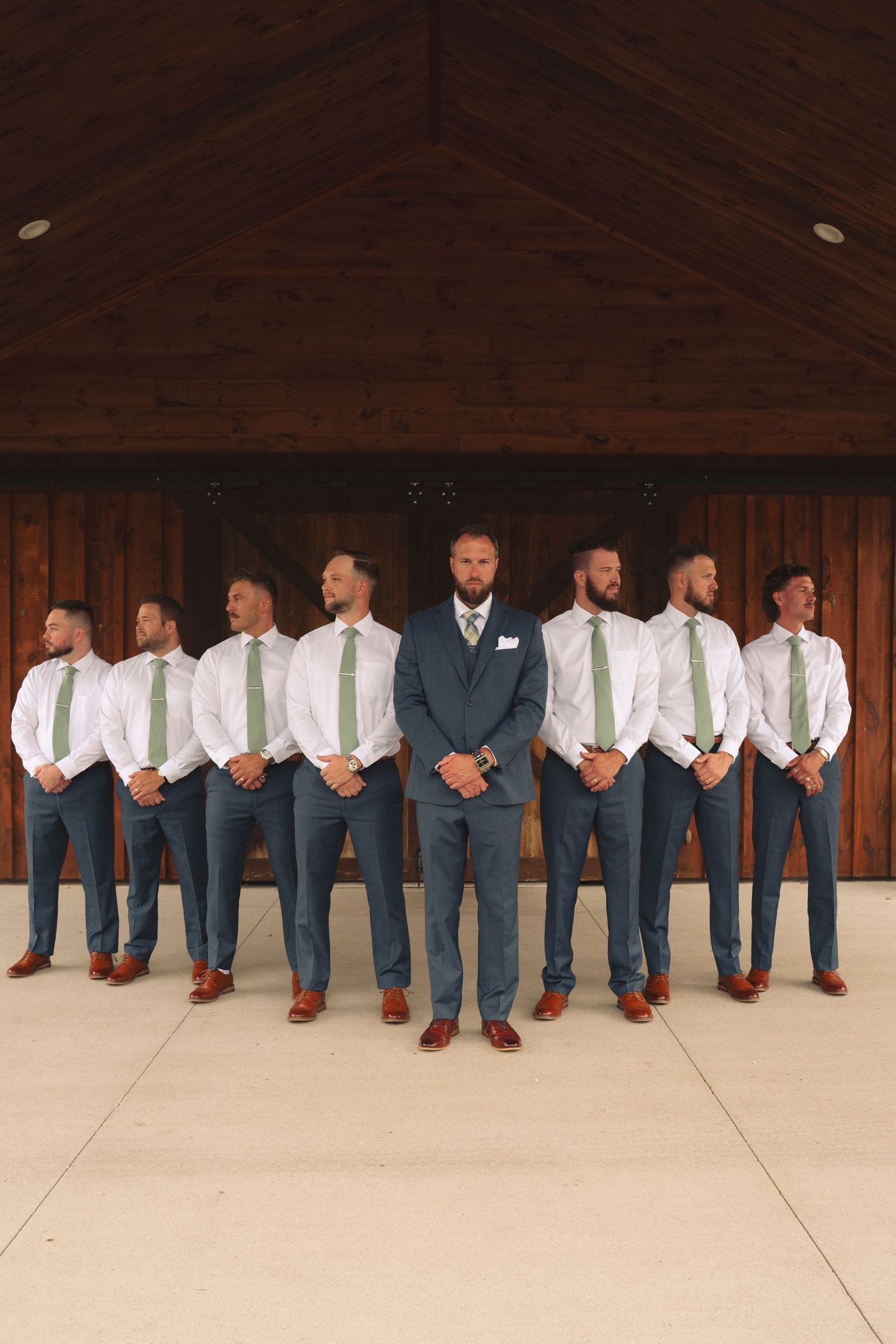 Groomsmen in navy suits and brown shoes lined up showing their coordinated footwear at a rustic wedding venue.
