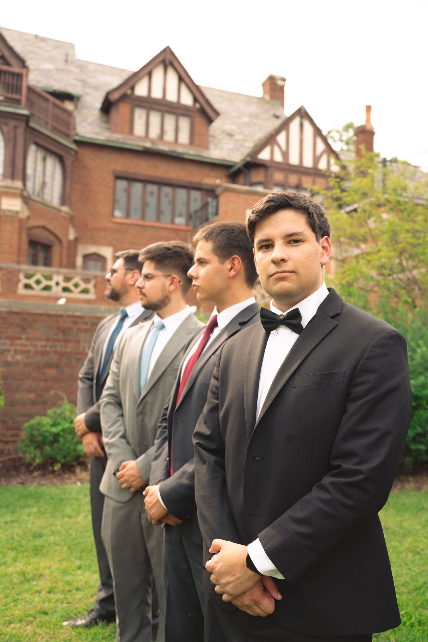 Groomsmen in formal attire stand in line outside historic brick building during wedding photo session.