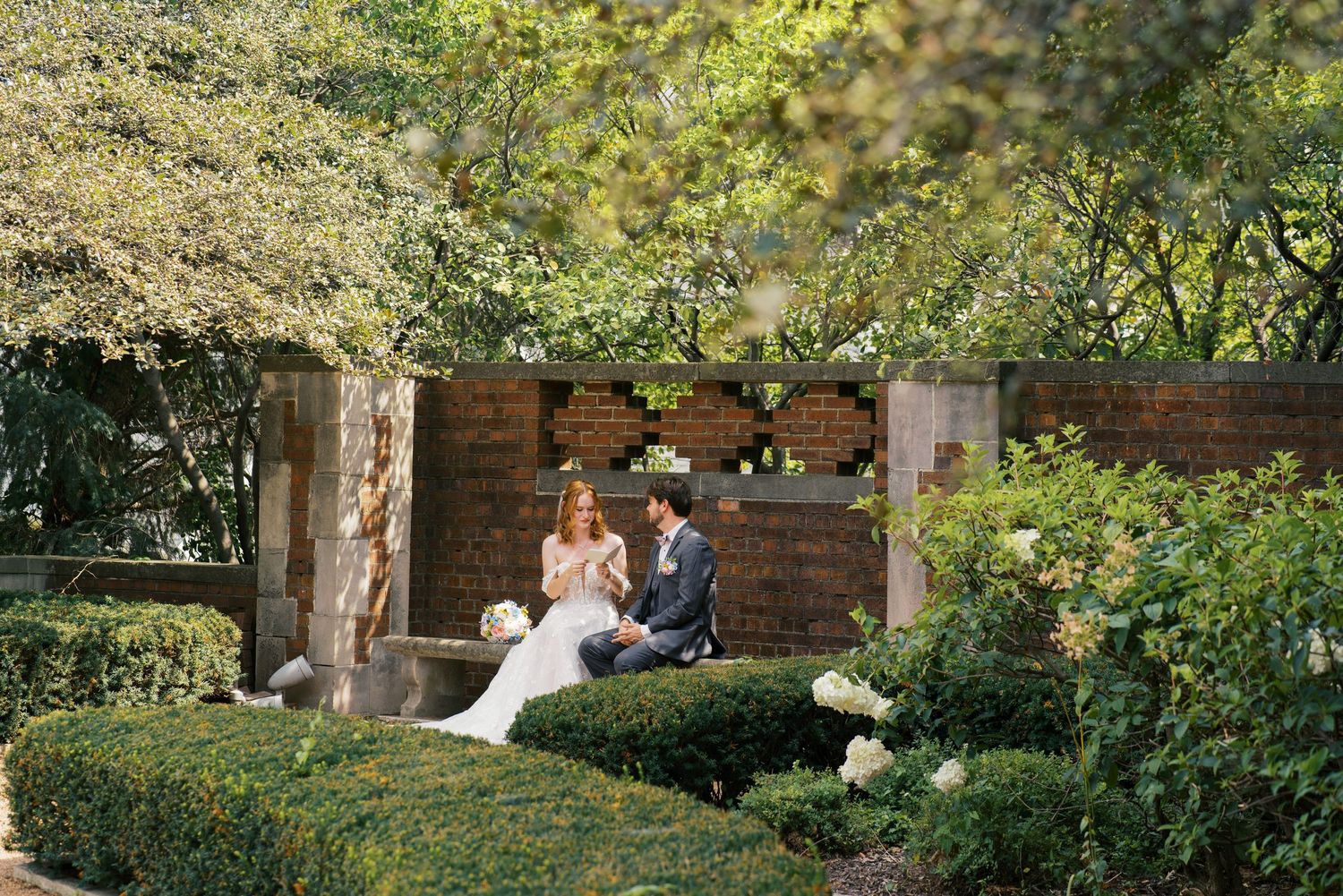 A bride and groom sit together in a lush garden surrounded by manicured hedges and a rustic brick wall backdrop.