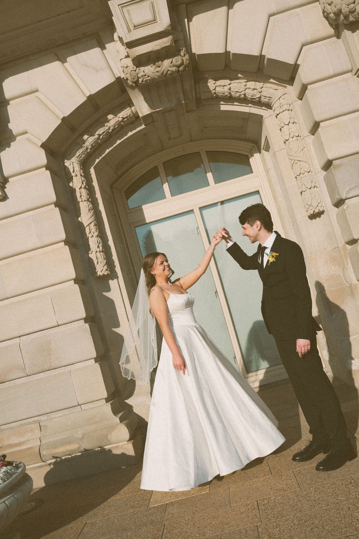 A bride and groom dance together outside an elegant stone building, she in a white gown and veil, he in a black tuxedo.