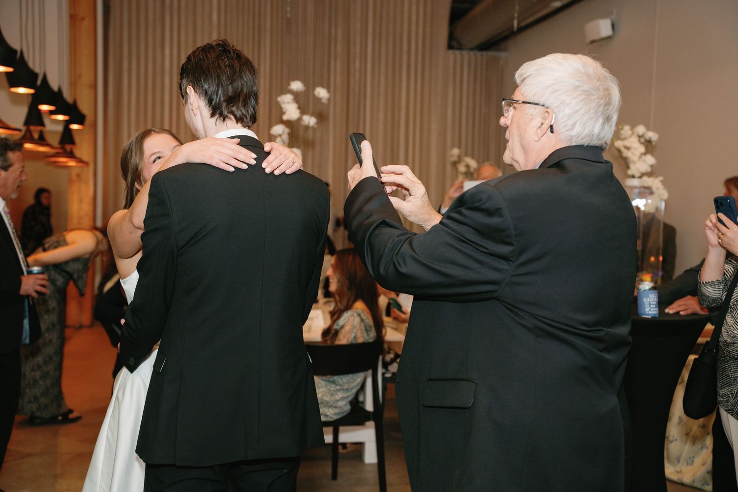 Elderly man photographs couple embracing at elegant wedding reception in modern venue.