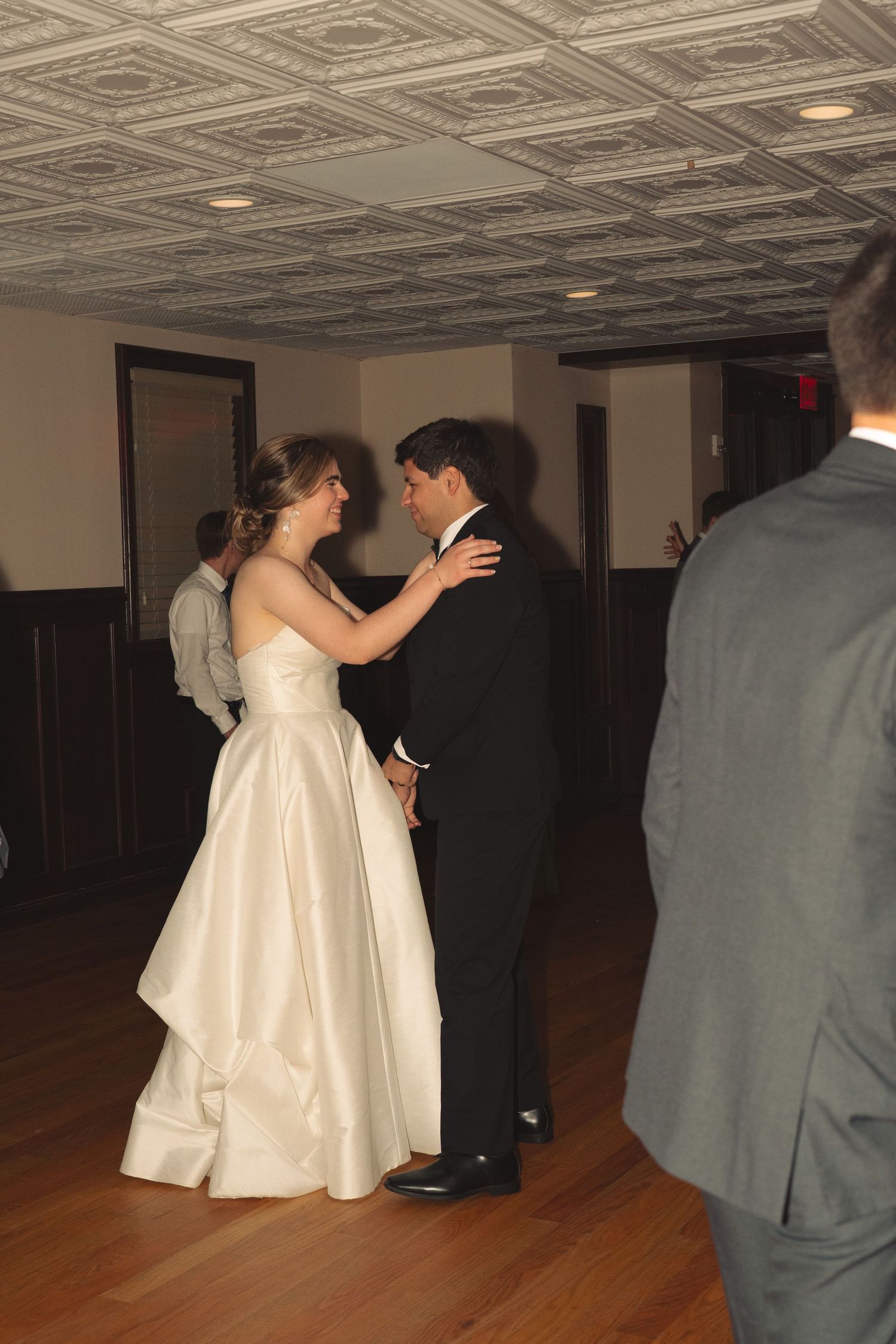 A couple in formal attire dances on a wooden floor beneath decorative ceiling tiles.