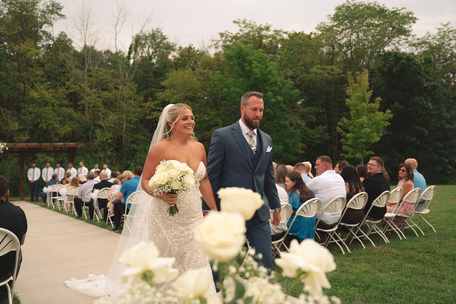 Panoramic view of wedding ceremony setup with guests seated in white chairs under trees.
