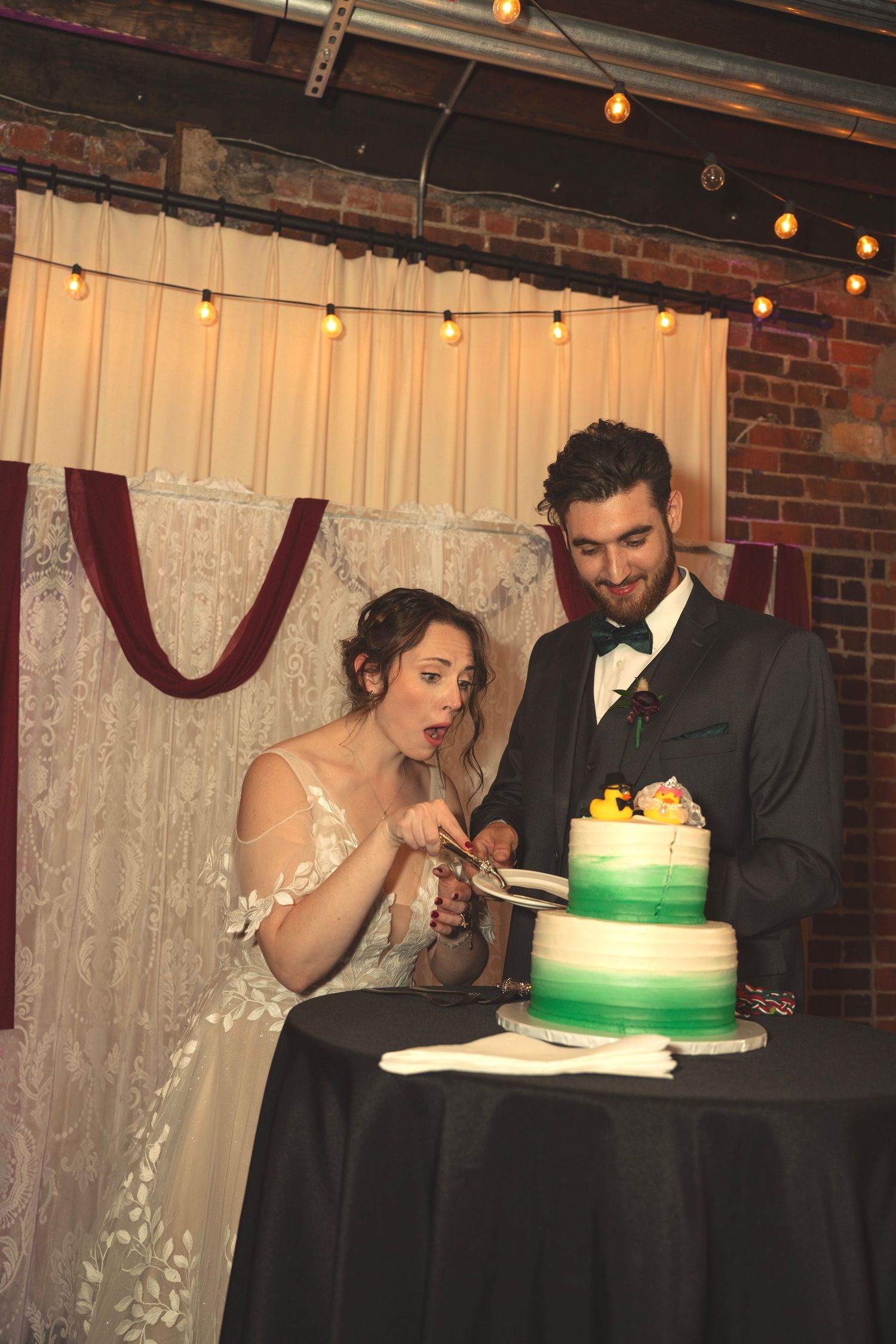 Couple enjoying their wedding cake with a unique green ombre design at a rustic brick venue.