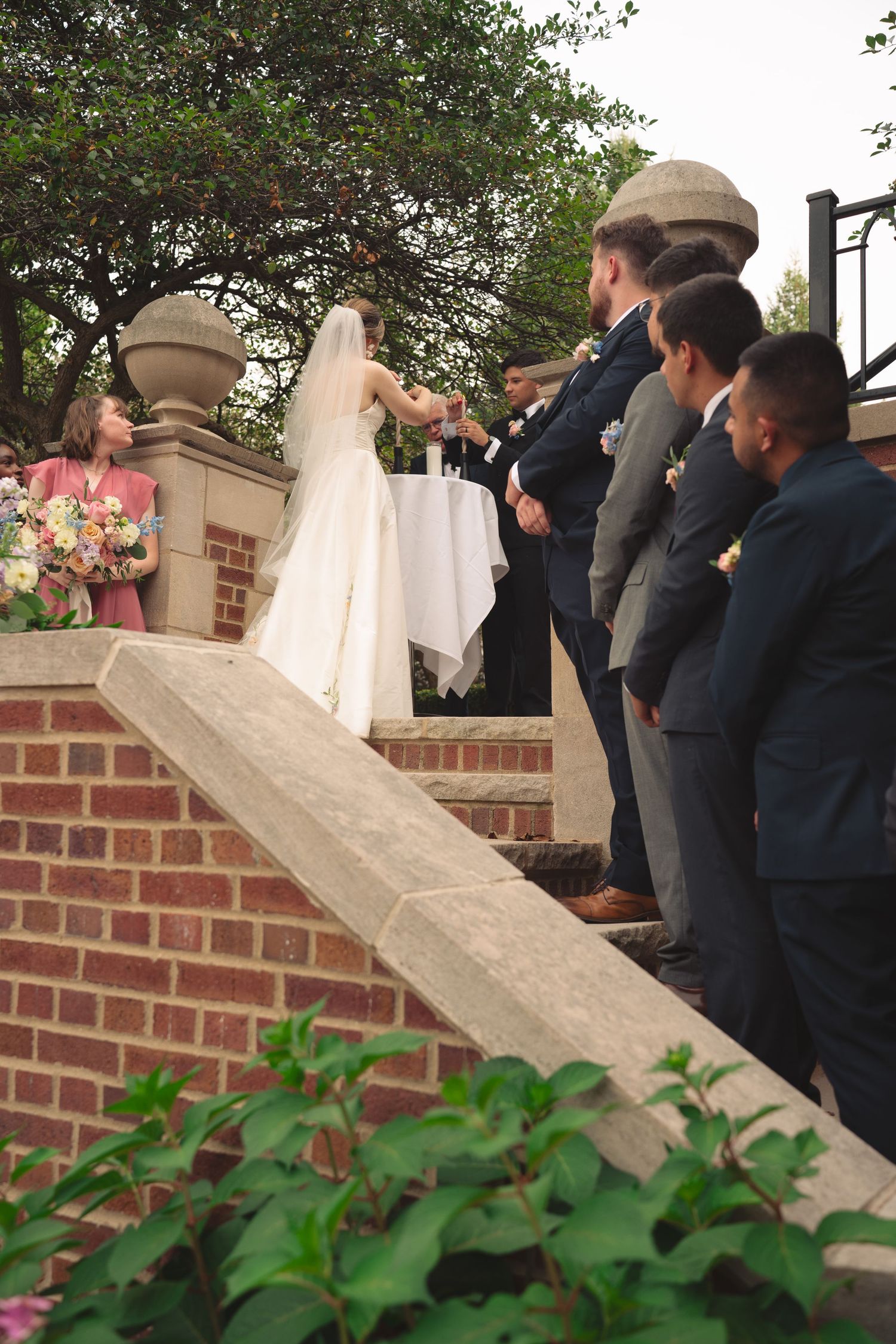 Wedding guests gather along a brick wall and staircase surrounded by flowering plants.
