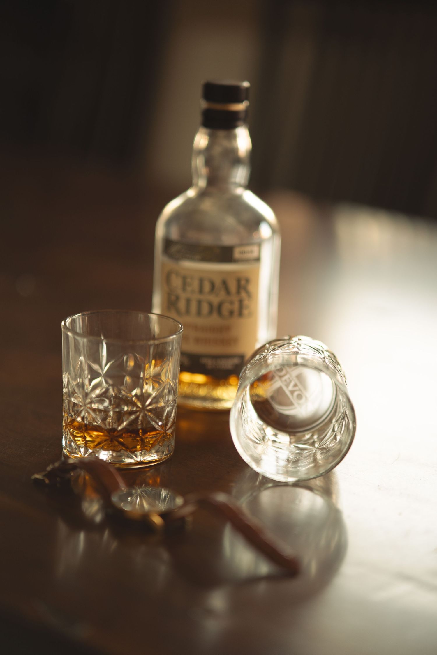 Crystal whiskey glasses and a bottle of Cedar Ridge bourbon on a dark wooden surface in dramatic lighting.
