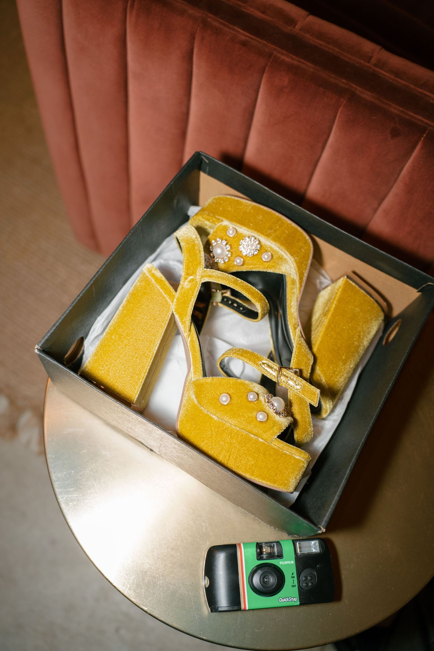 Yellow velvet platform heels with pearl embellishments in a box beside a disposable camera on a gold table.