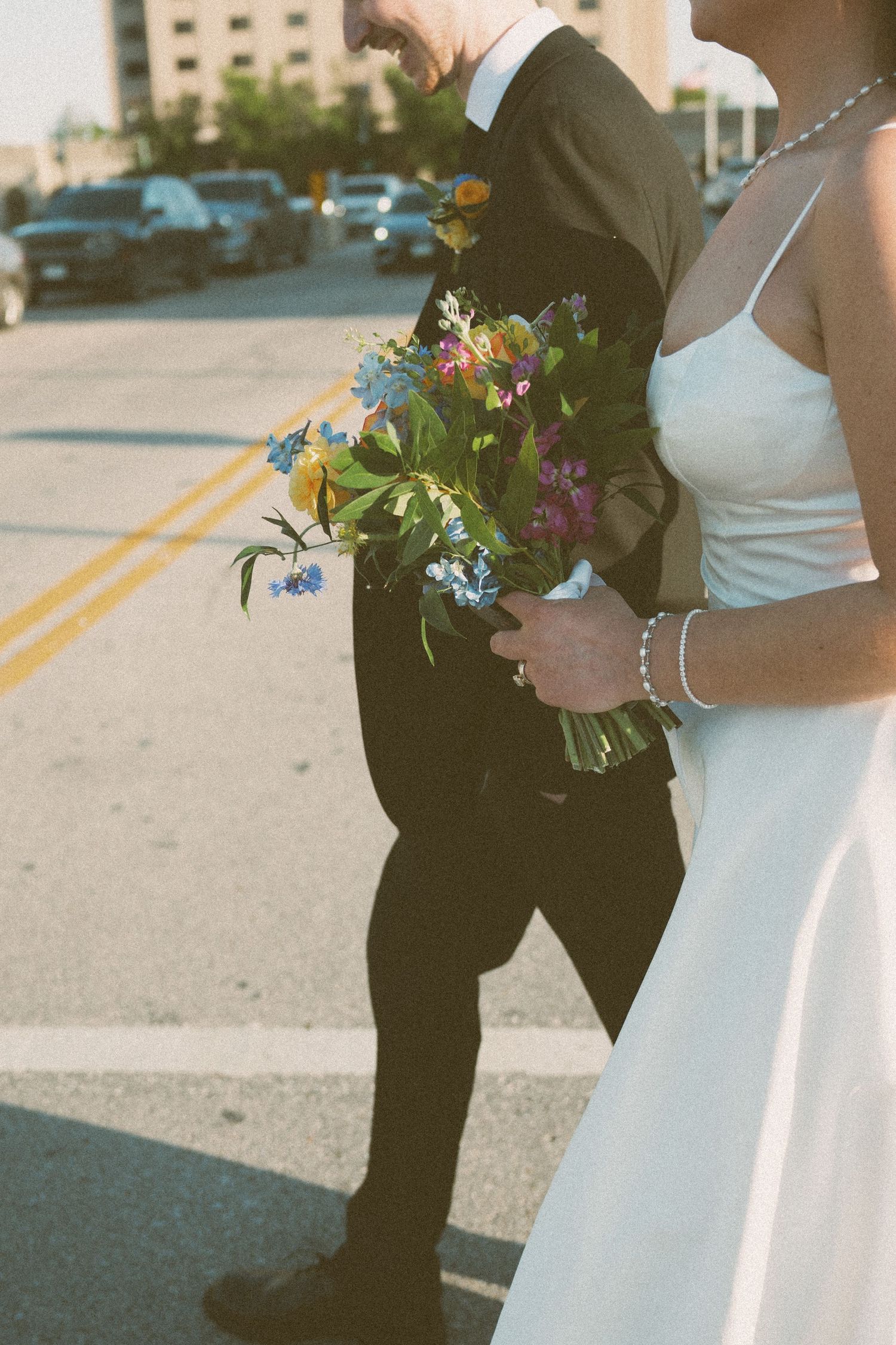 Bride and groom walking across urban street, bride holding colorful wildflower bouquet, wearing white dress and black suit.
