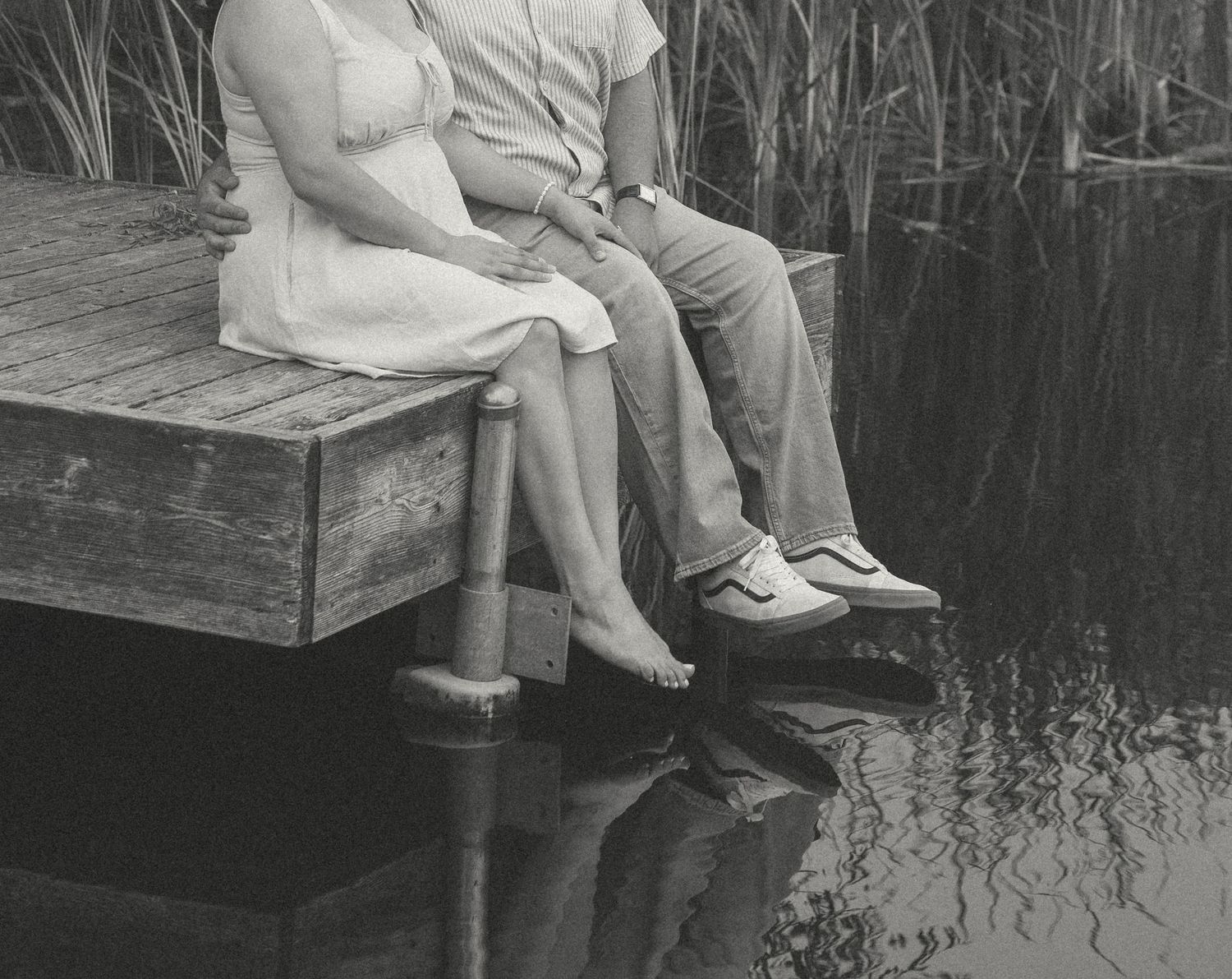 Couple sitting together on a wooden dock over calm water, surrounded by tall reeds, in a black and white photo.