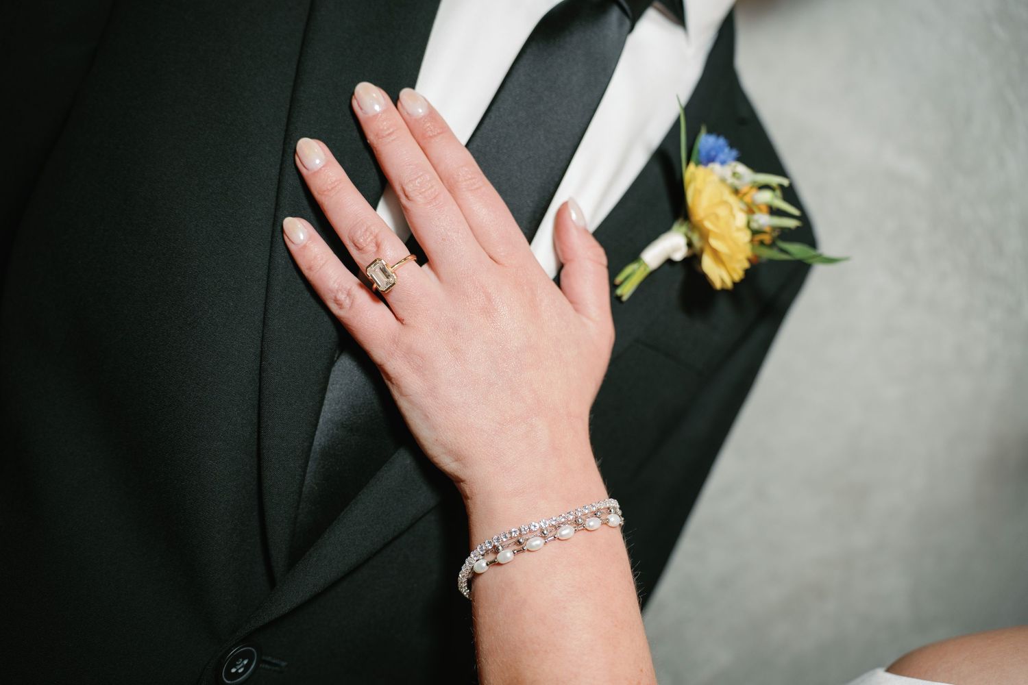 Bride's hand with diamond ring and bracelet resting on groom's black tuxedo lapel with yellow boutonniere.