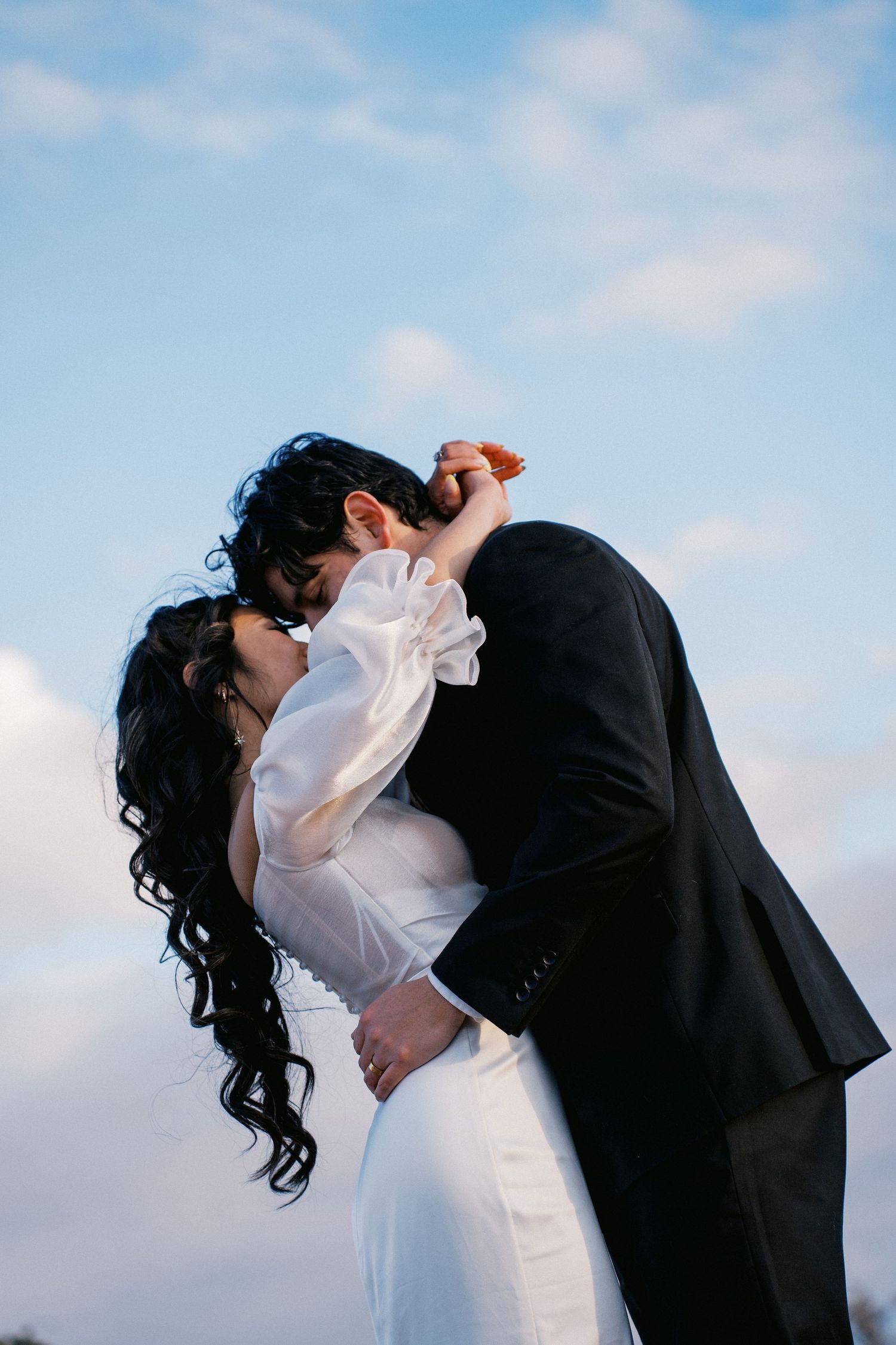 A bride and groom share a romantic kiss against a blue sky, she in white dress, he in black suit.