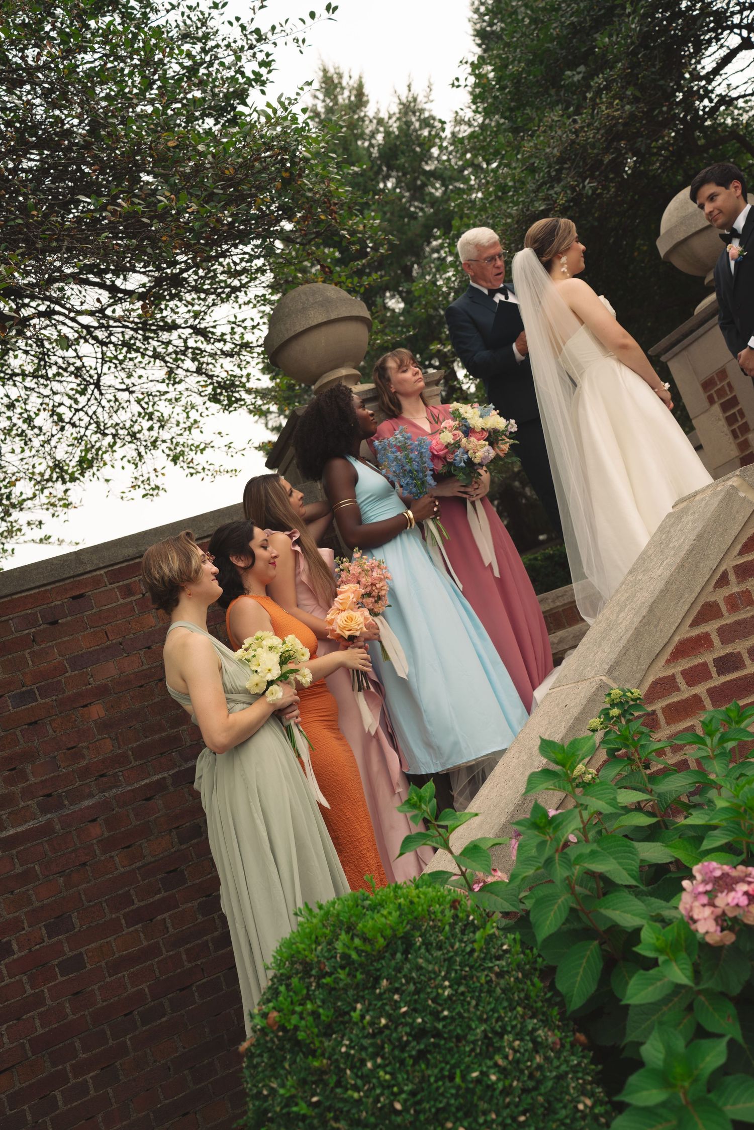 Wedding party members in pastel dresses gather near flowering bushes and brick walls.