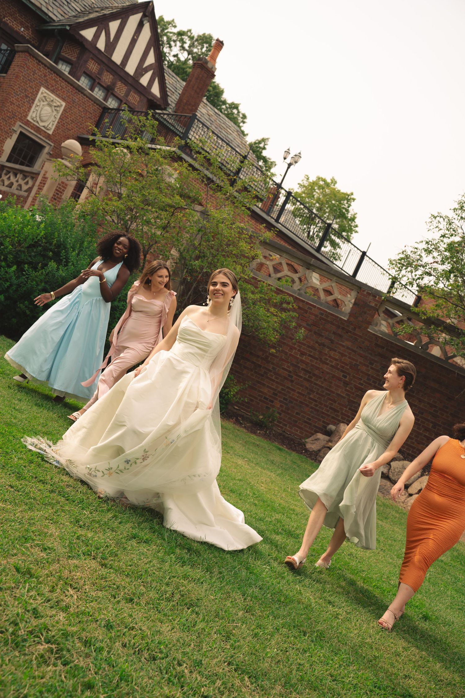 Wedding party members in light dresses twirl and dance playfully on a grassy lawn outside a Tudor-style brick building.