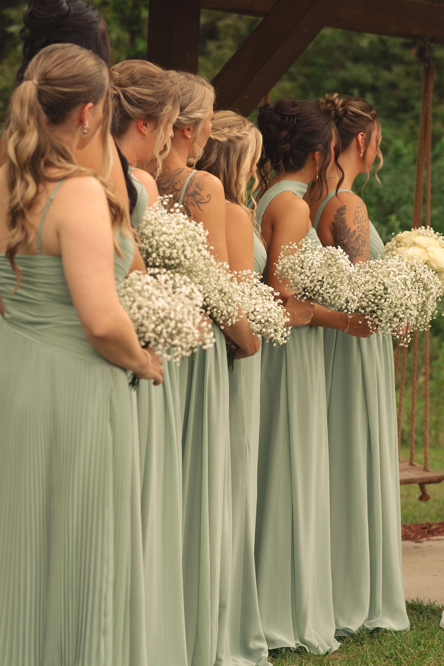 Bridesmaids in sage green dresses hold baby's breath bouquets while standing in a row at an outdoor wedding ceremony.