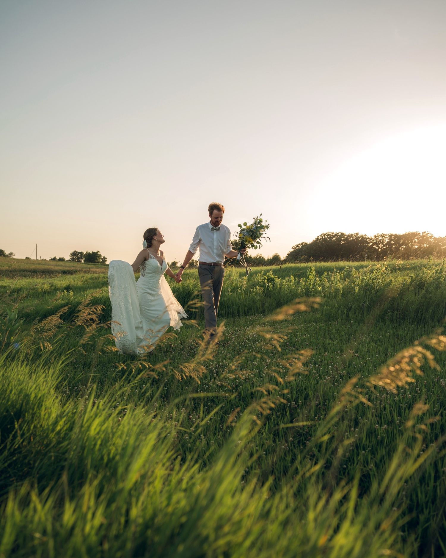 Couple walking hand in hand through tall grass during golden hour sunset.