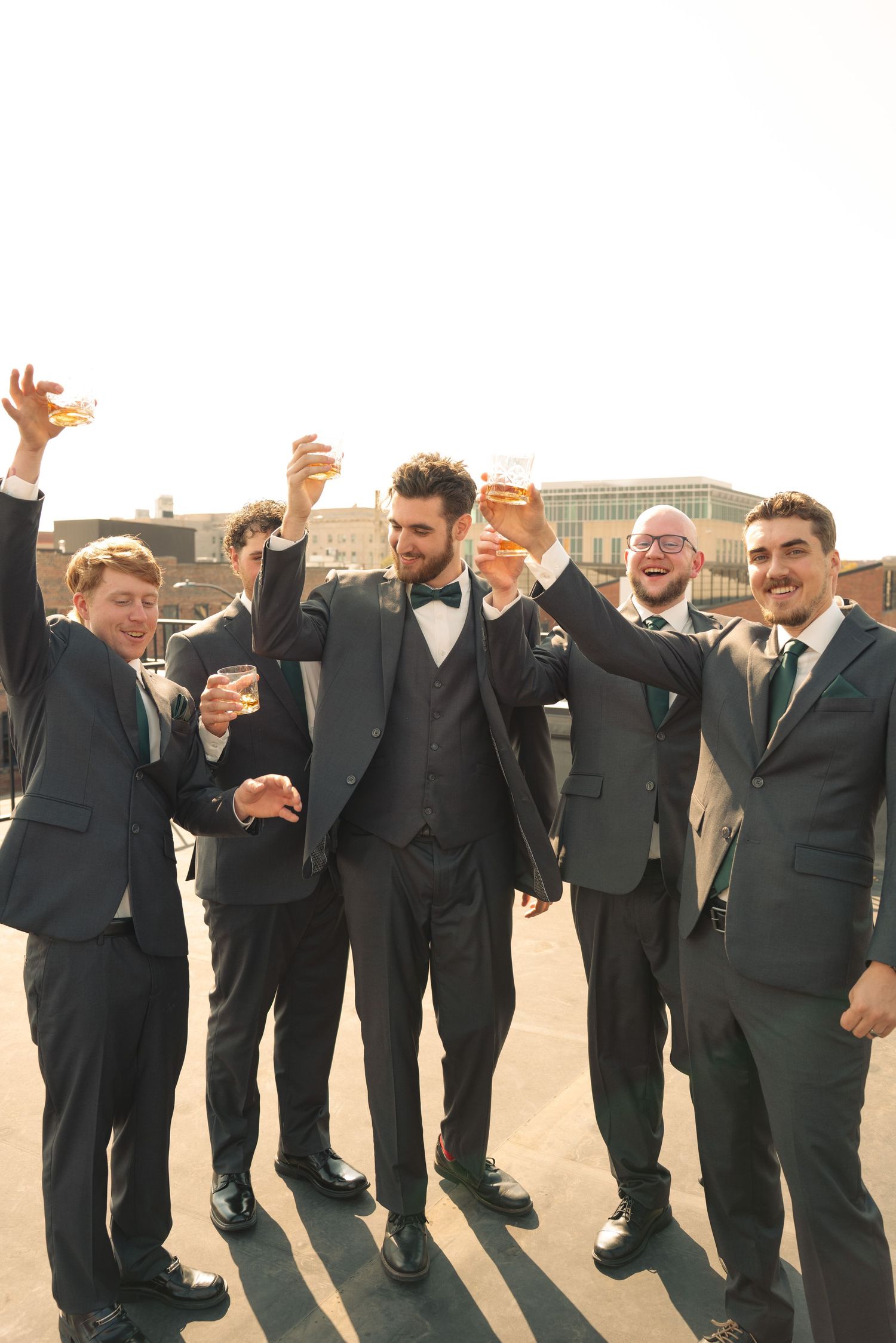 Groomsmen in black suits pose playfully in a line outdoors.
