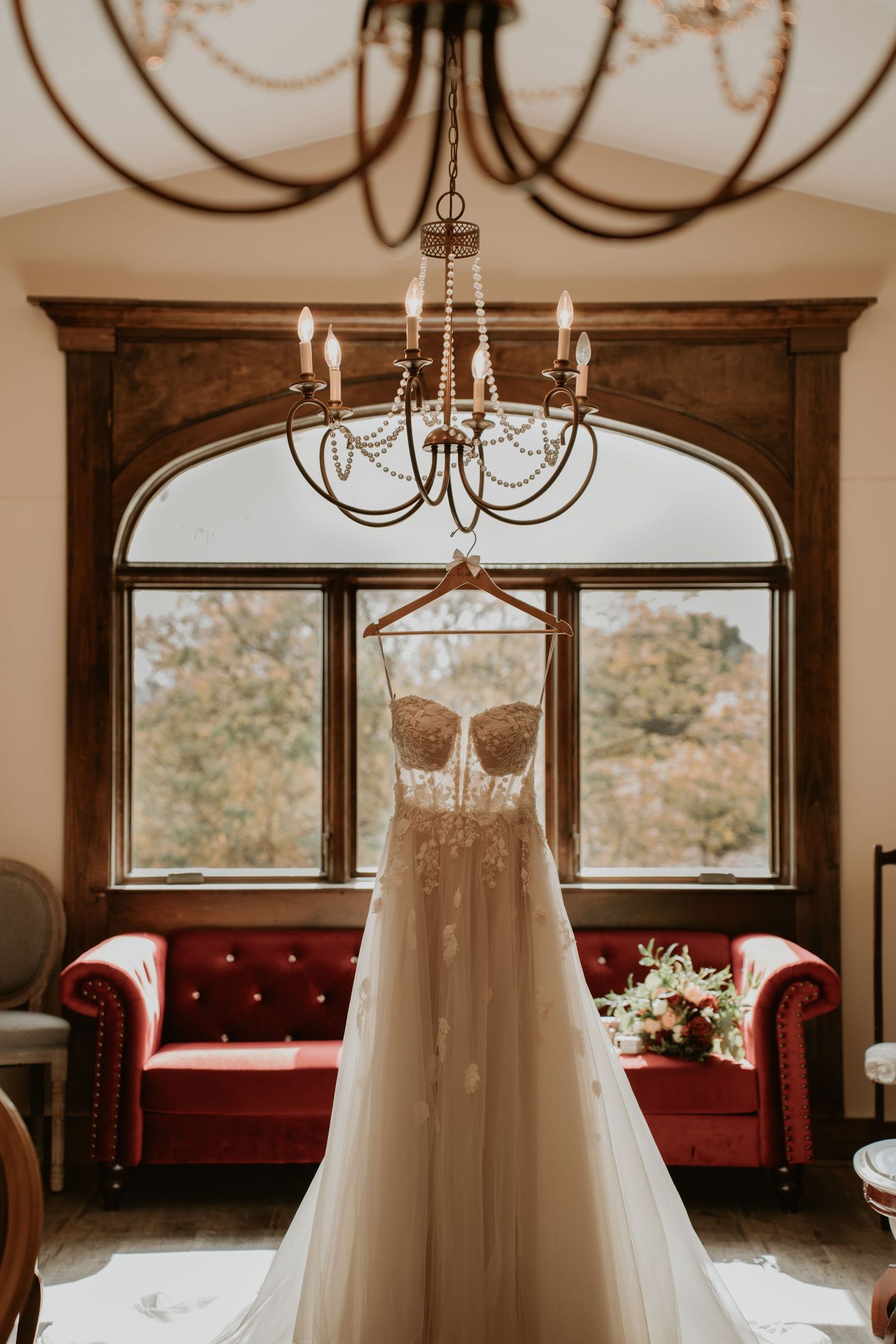 White wedding dress hangs elegantly from chandelier in sunlit room with arched windows and red velvet couch.