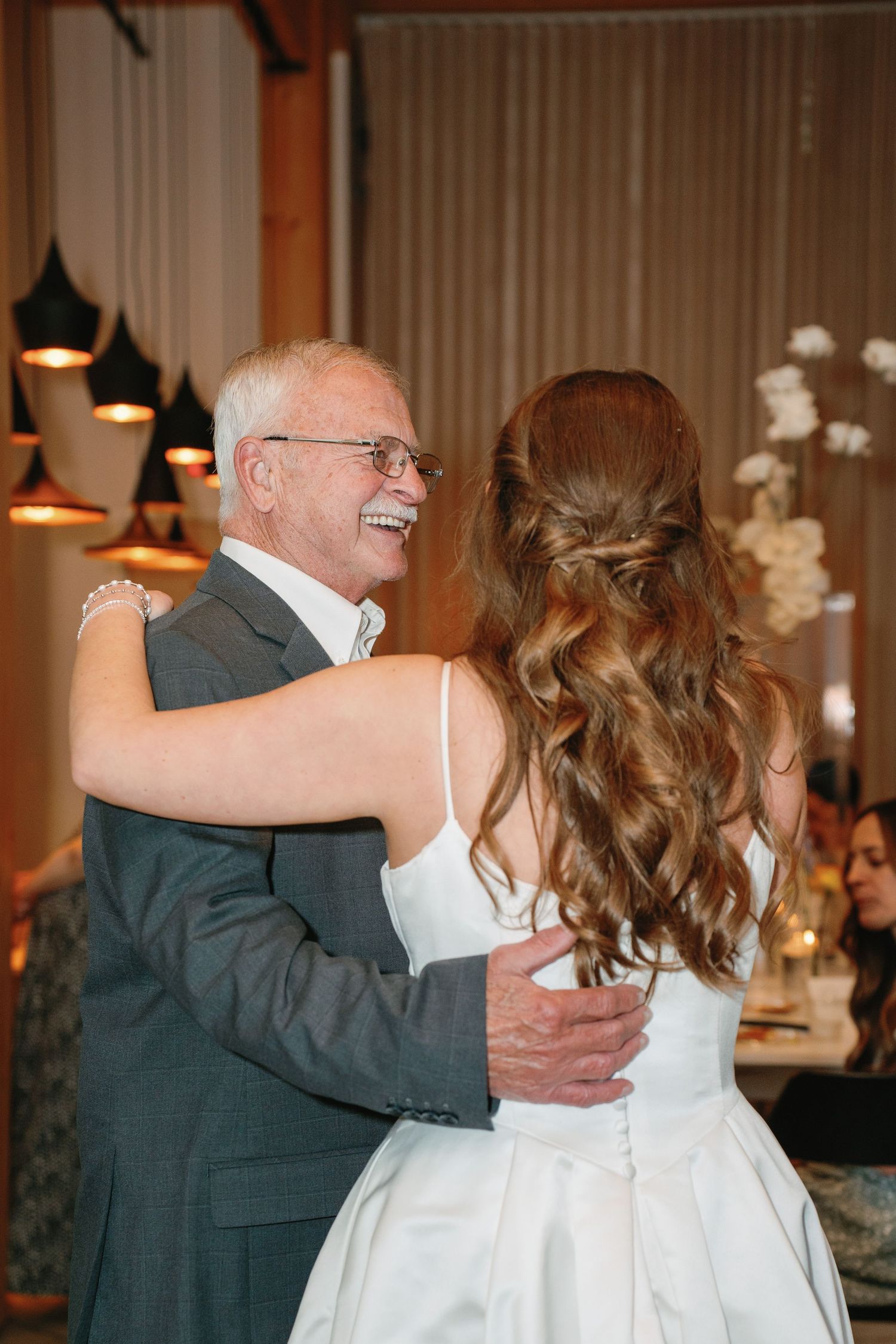 Father-daughter wedding dance with bride in white gown and older man in gray suit sharing a joyful moment.