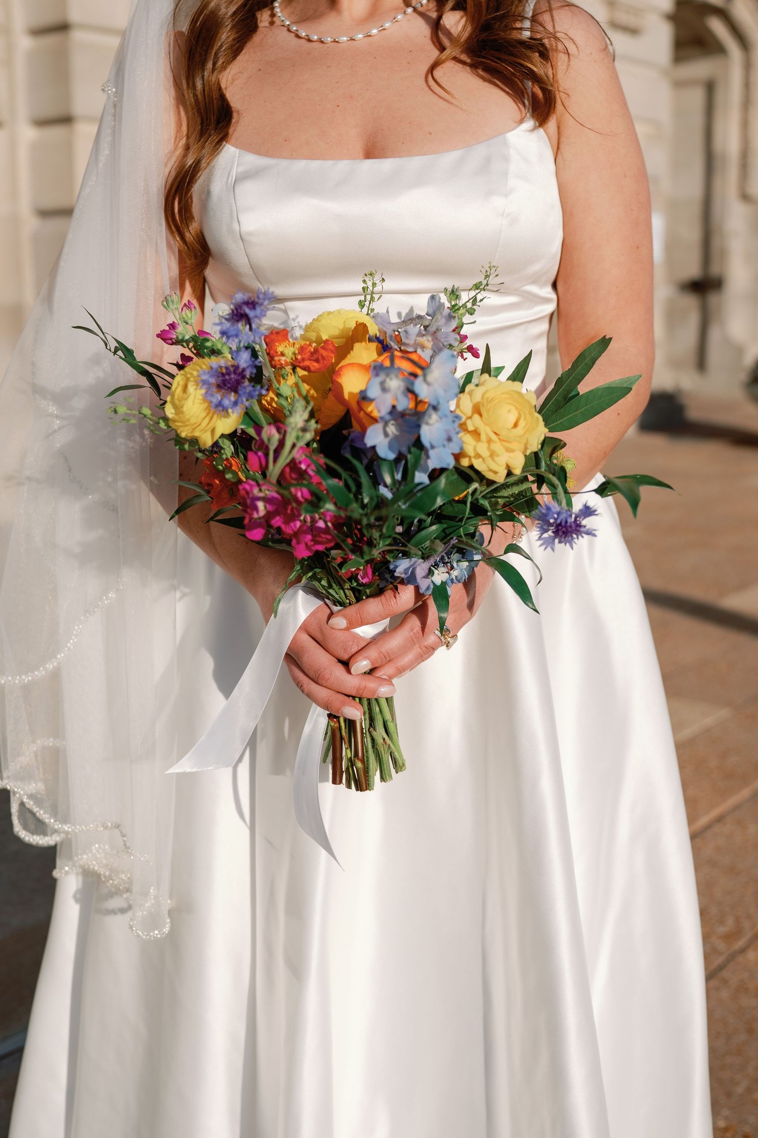 Bride in white satin gown holding colorful wedding bouquet with yellow, blue, and purple flowers tied with white ribbon.