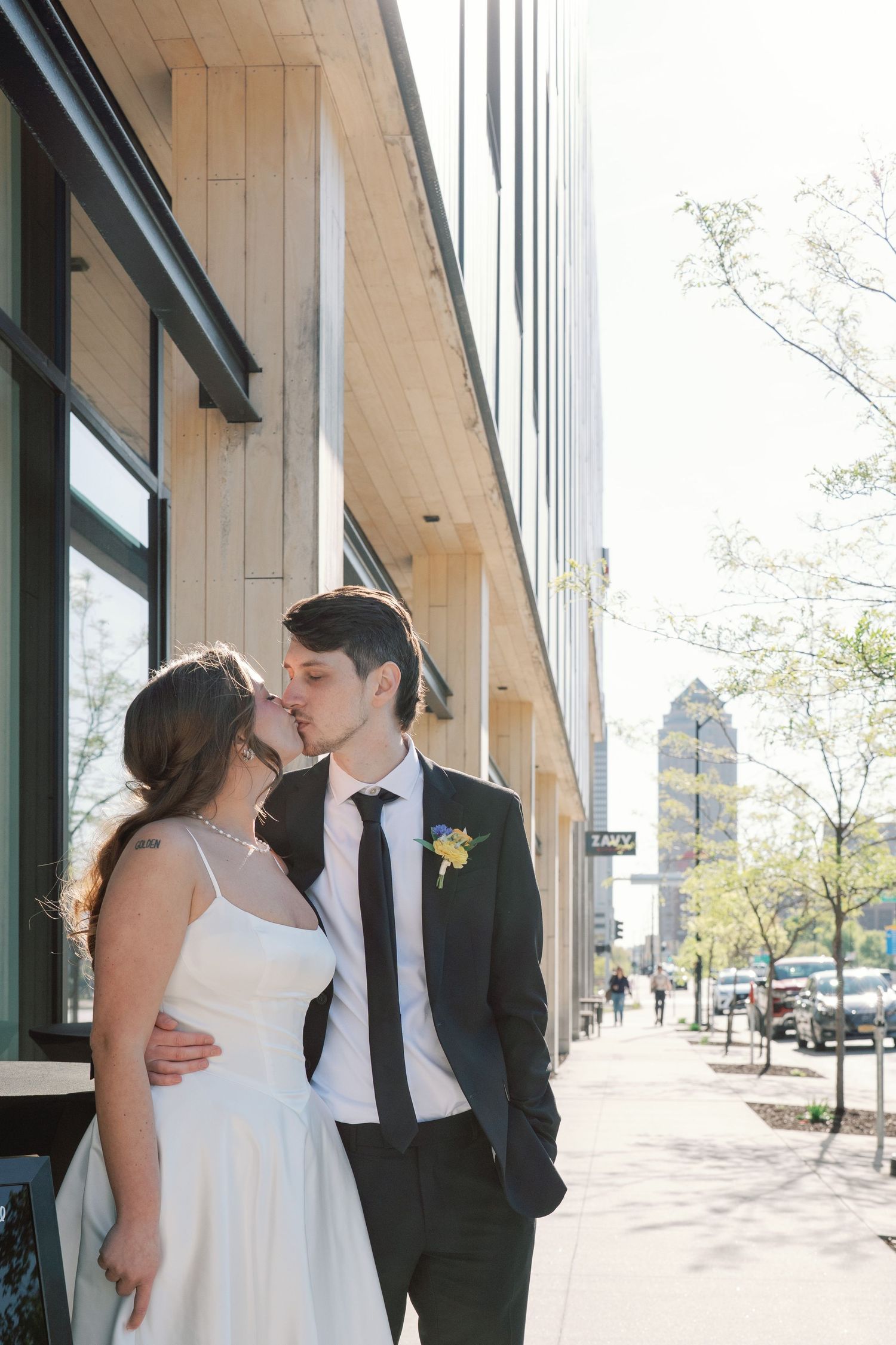 A bride and groom share a kiss on a sunny urban sidewalk, with the groom in a dark suit and boutonniere.