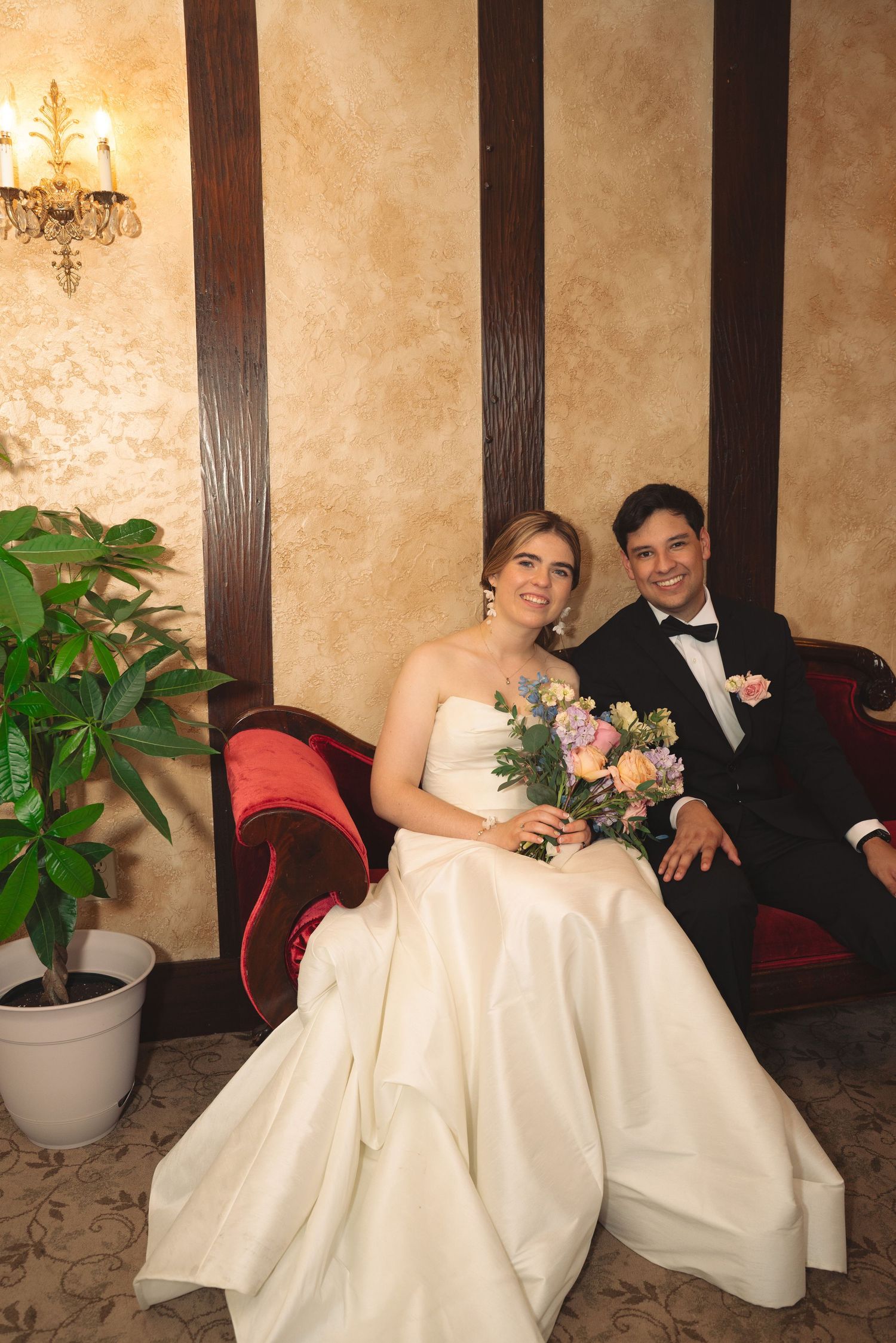 Elegant wedding couple seated on red velvet couch with floral bouquet in ornate interior.