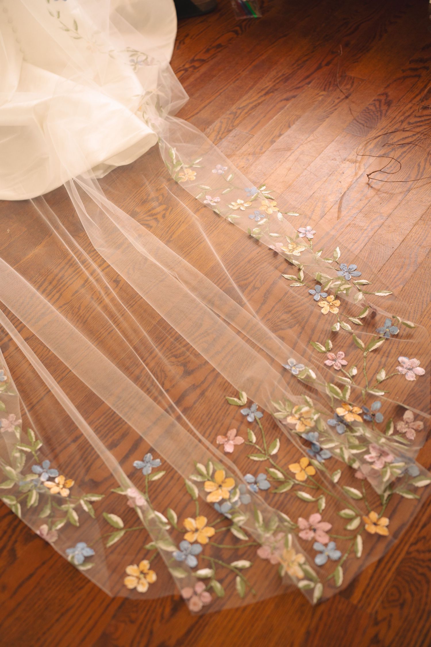 Close-up of colorful floral embroidery on white wedding veil hem against hardwood flooring.