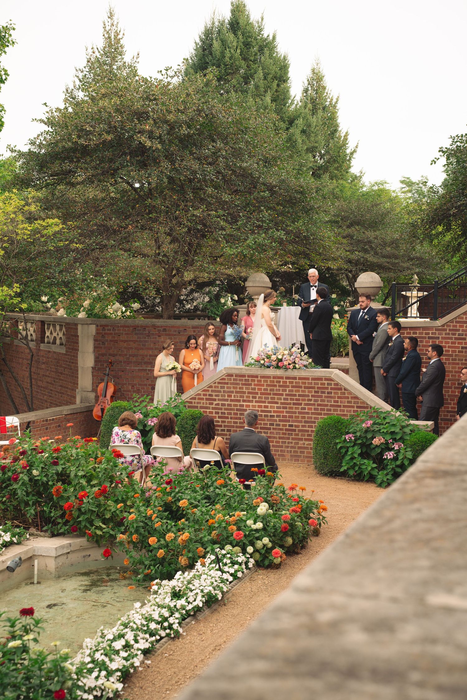 Wedding ceremony setup along a brick walkway surrounded by lush flower gardens and hedges.