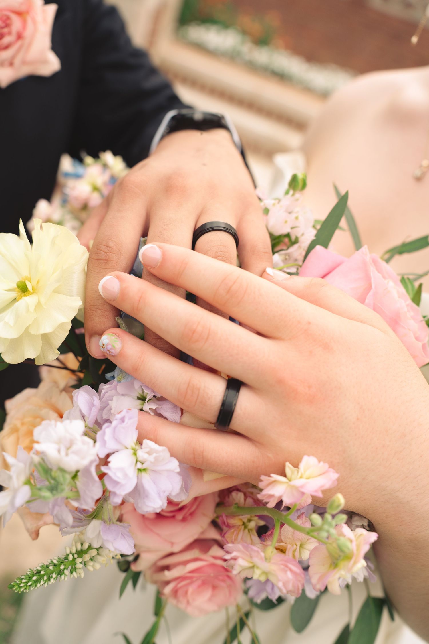Close-up of hands wearing black wedding bands resting on pink and purple floral bouquet.