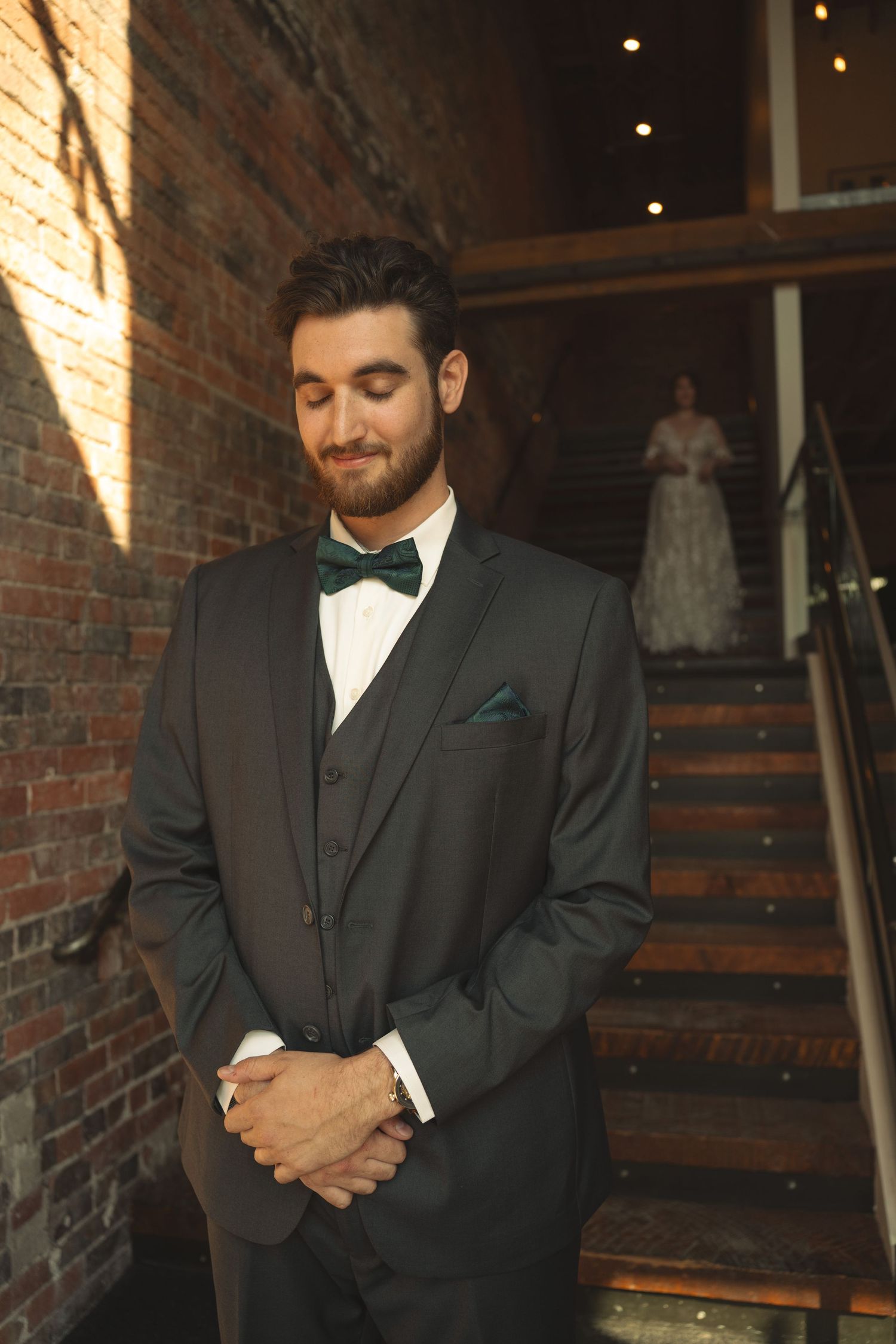 Groom in dark tuxedo with bow tie poses thoughtfully in front of rustic brick wall.
