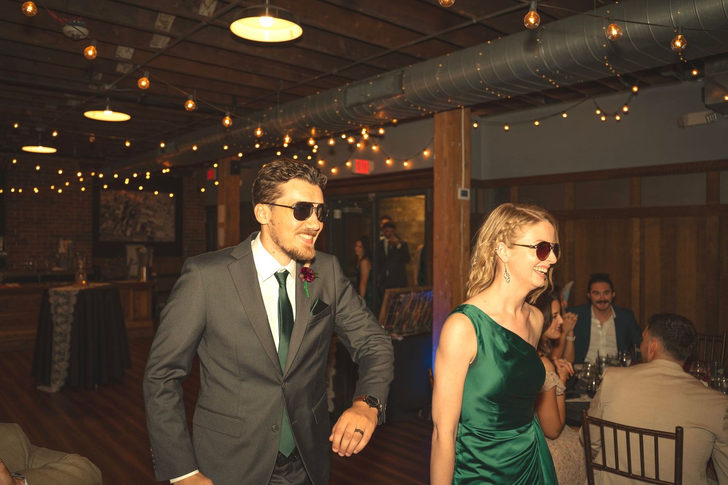 Wedding party dancing in sunglasses under string lights at indoor reception.
