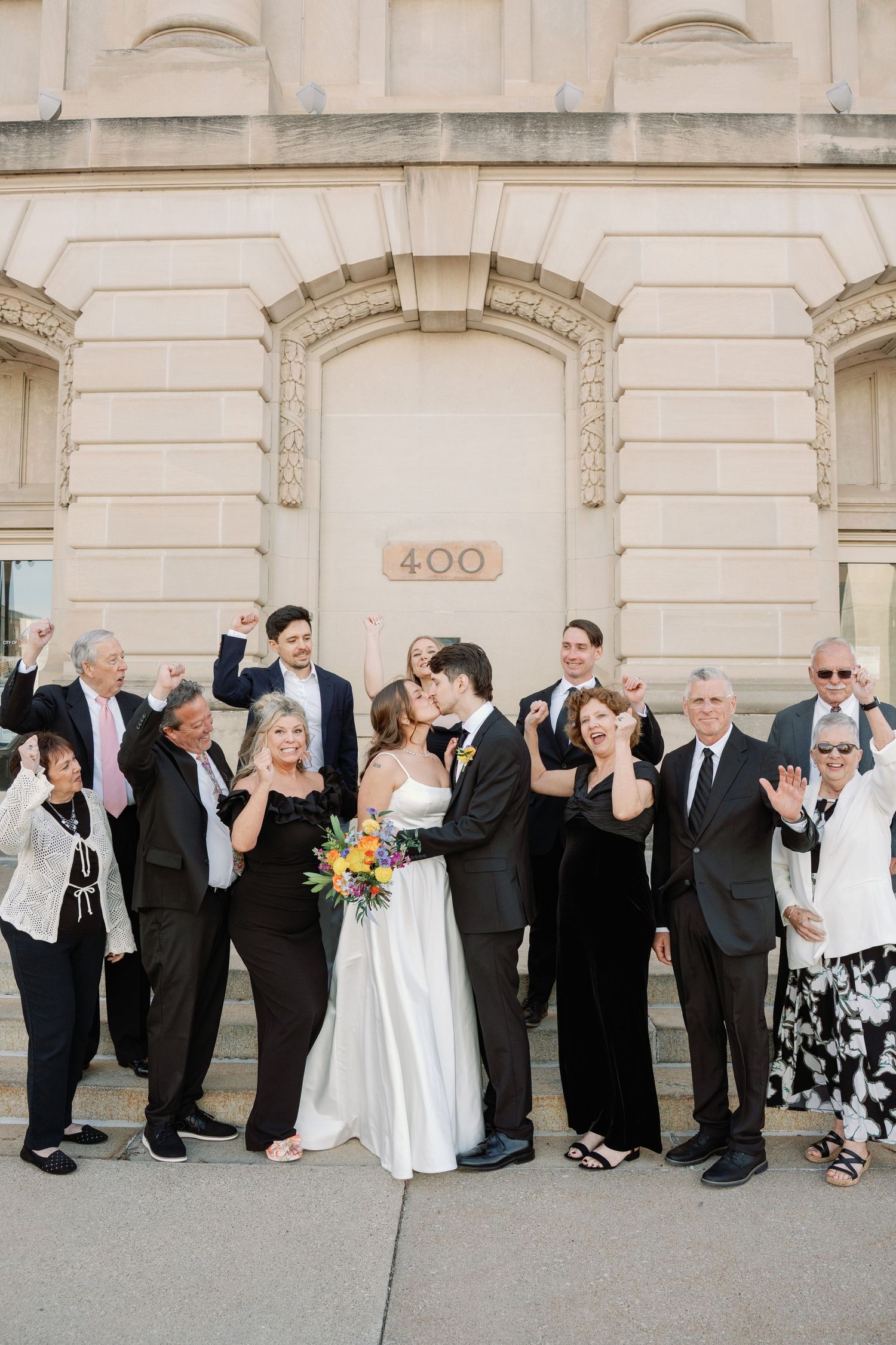 A bride and groom share a kiss surrounded by cheering family and friends outside a classic stone building numbered 400.