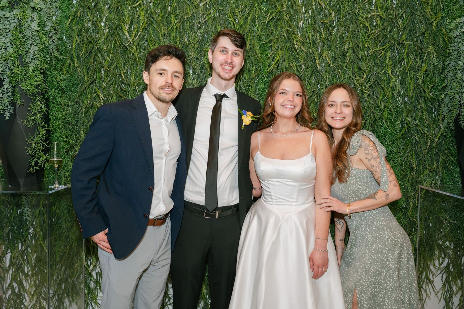 Two men and two women posing together at a wedding reception in front of a lush green leafy wall backdrop.