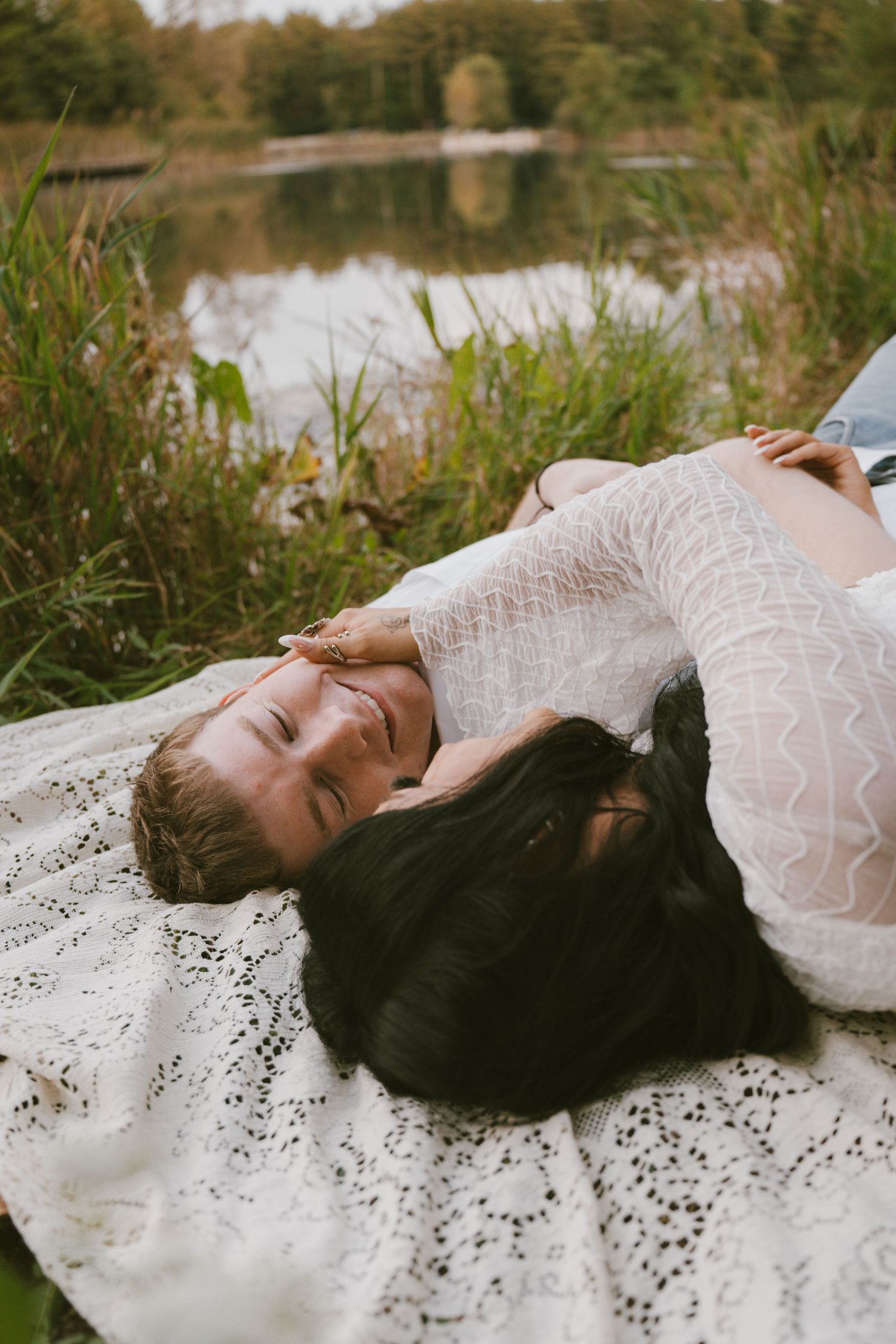 Pregnant woman in white lace dress lying on blanket by a serene lake, smiling peacefully outdoors.