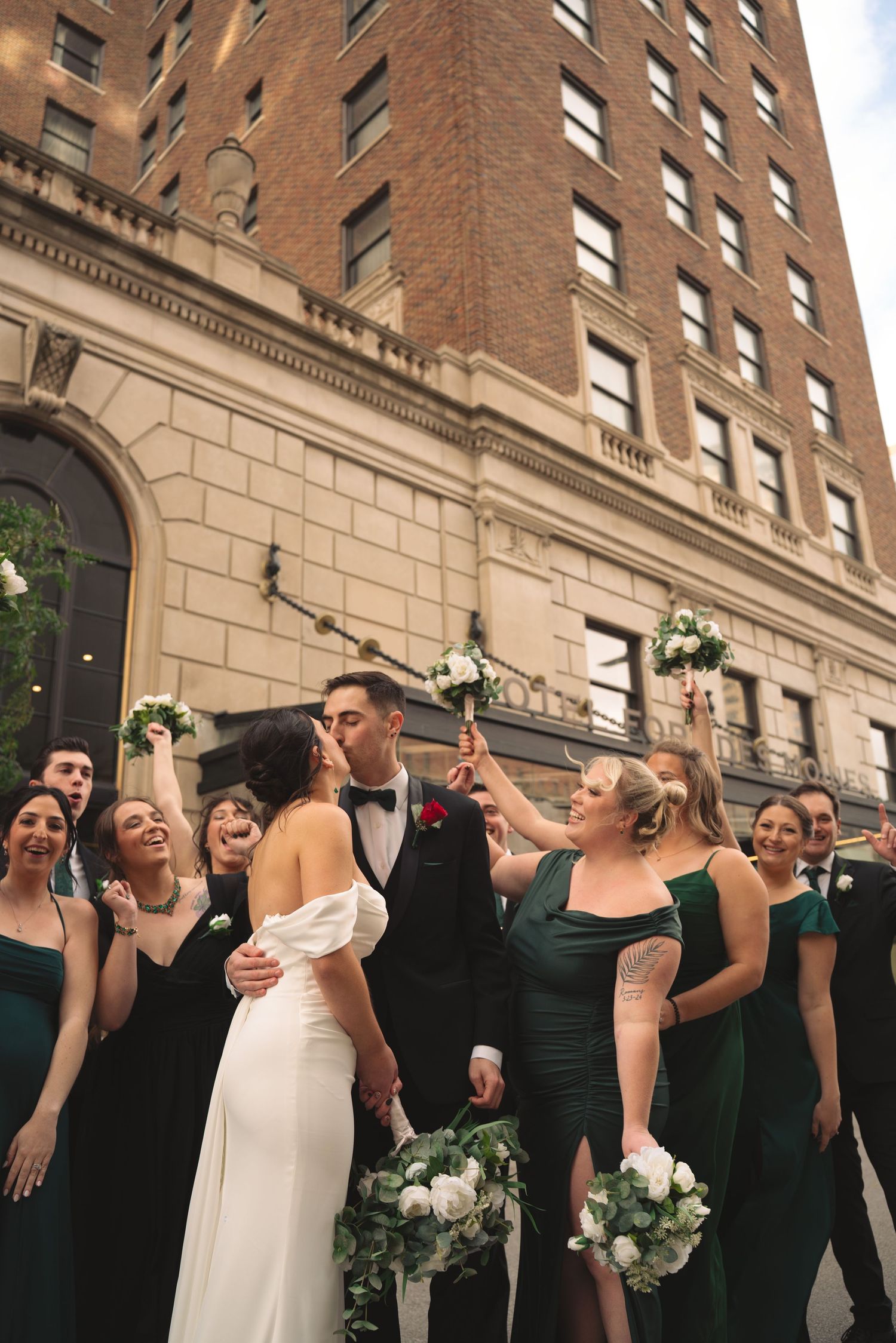 A wedding party in dark green dresses and white gowns poses together in front of a brick building.