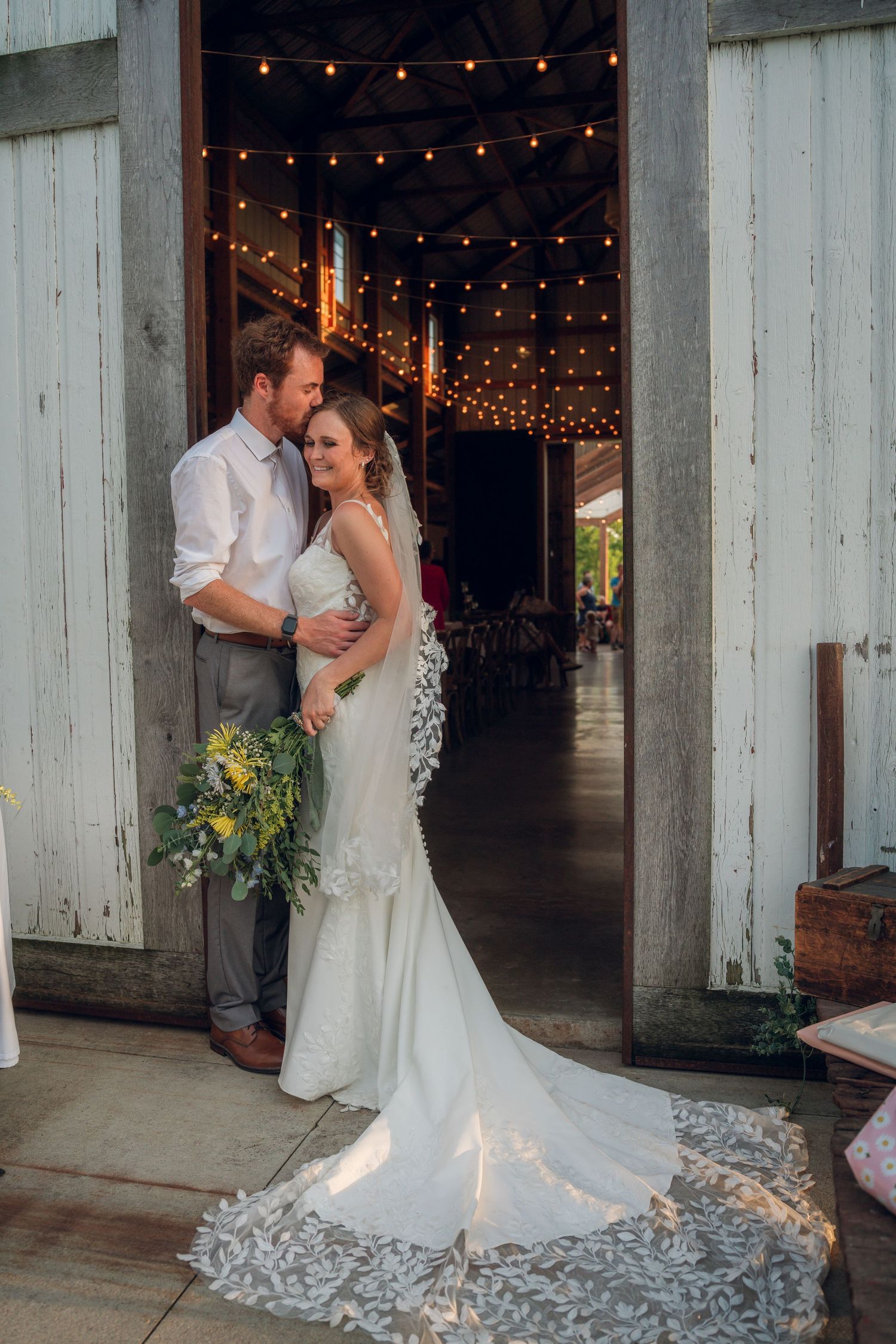 Wedding couple embraces under string lights in rustic barn setting.