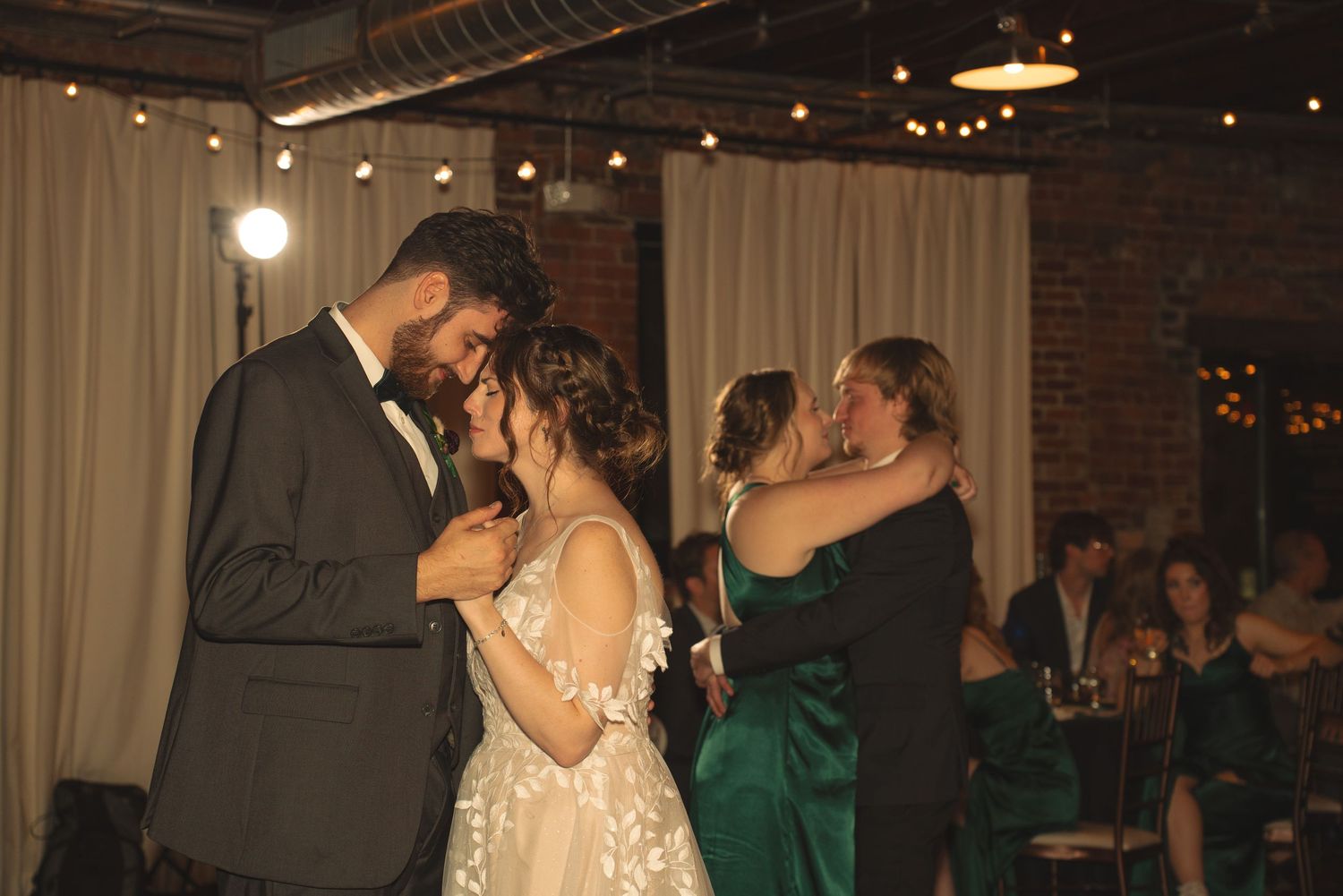 Wedding reception dance floor scene in a rustic brick-walled venue with string lights.