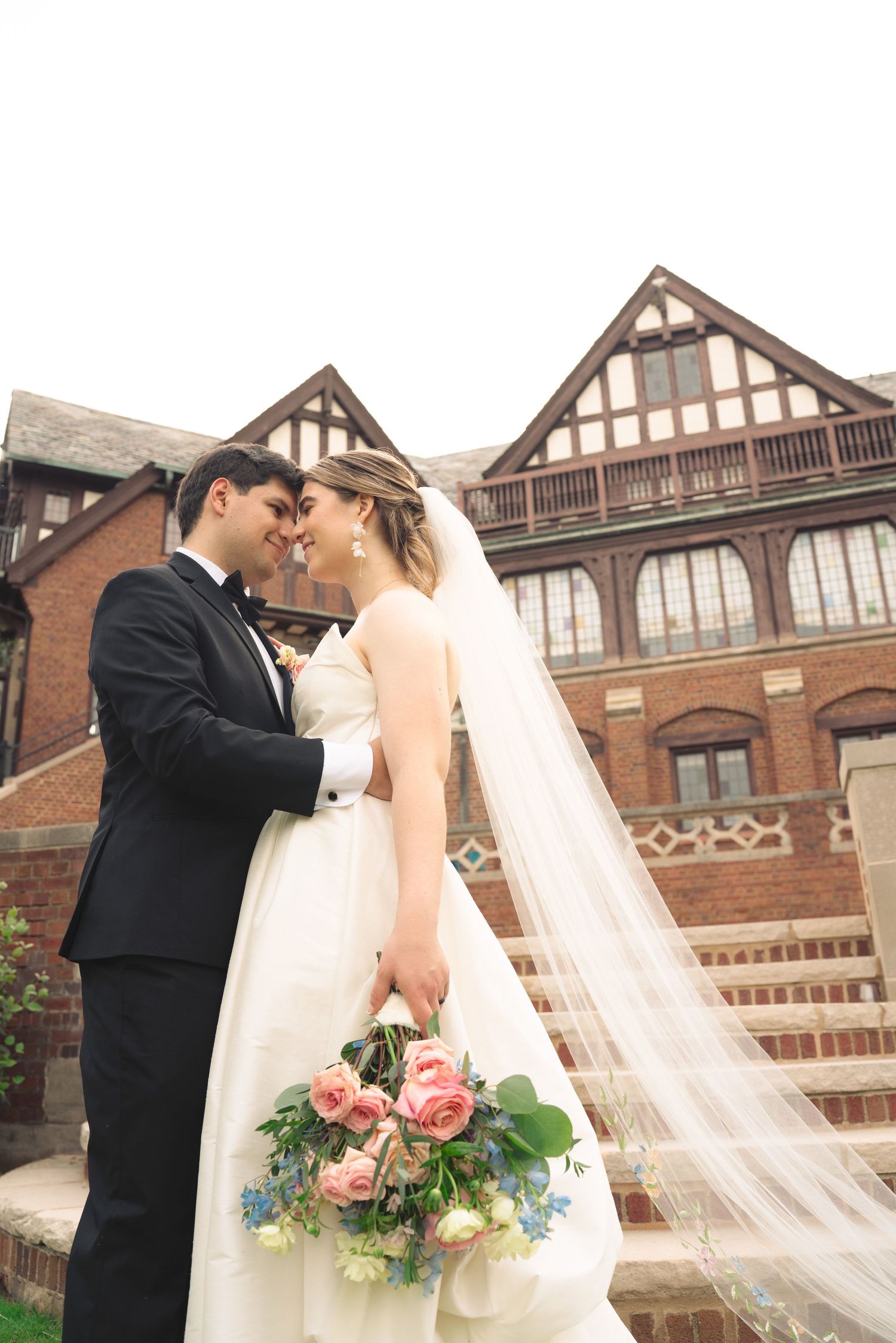 Romantic wedding portrait in front of Tudor-style architecture with flowing veil and bouquet.