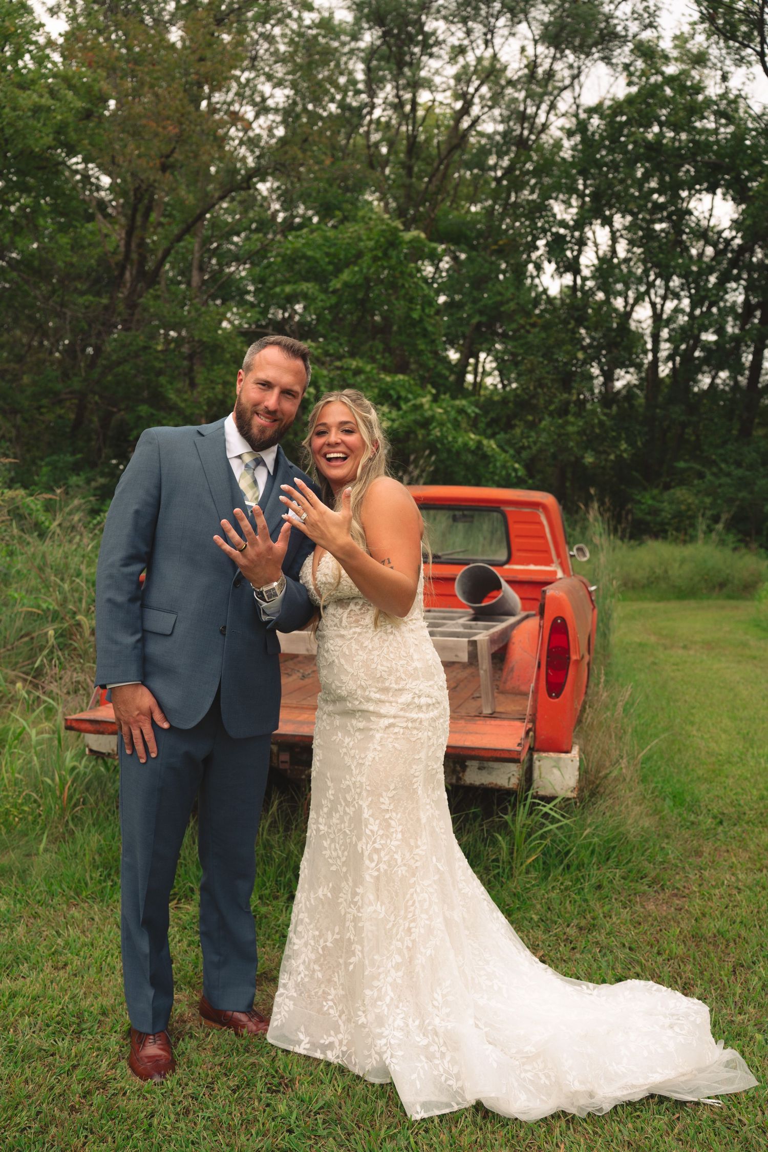Wedding couple posing playfully by vintage red pickup truck in grassy field.