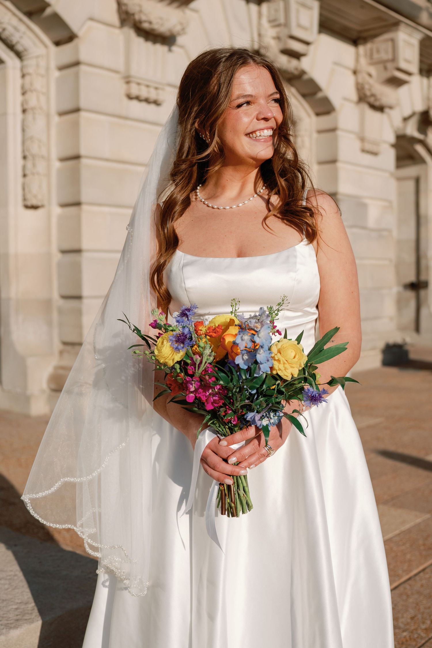 Smiling bride in white satin gown and veil holds colorful bouquet outside elegant stone building.