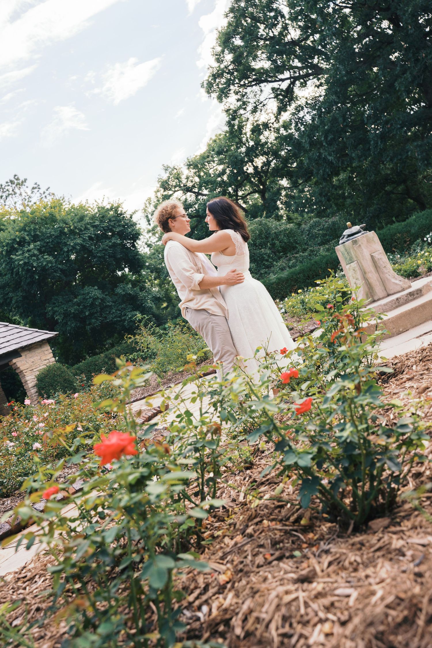 Couple embracing romantically in a lush garden surrounded by blooming red roses and green trees on a cloudy day.