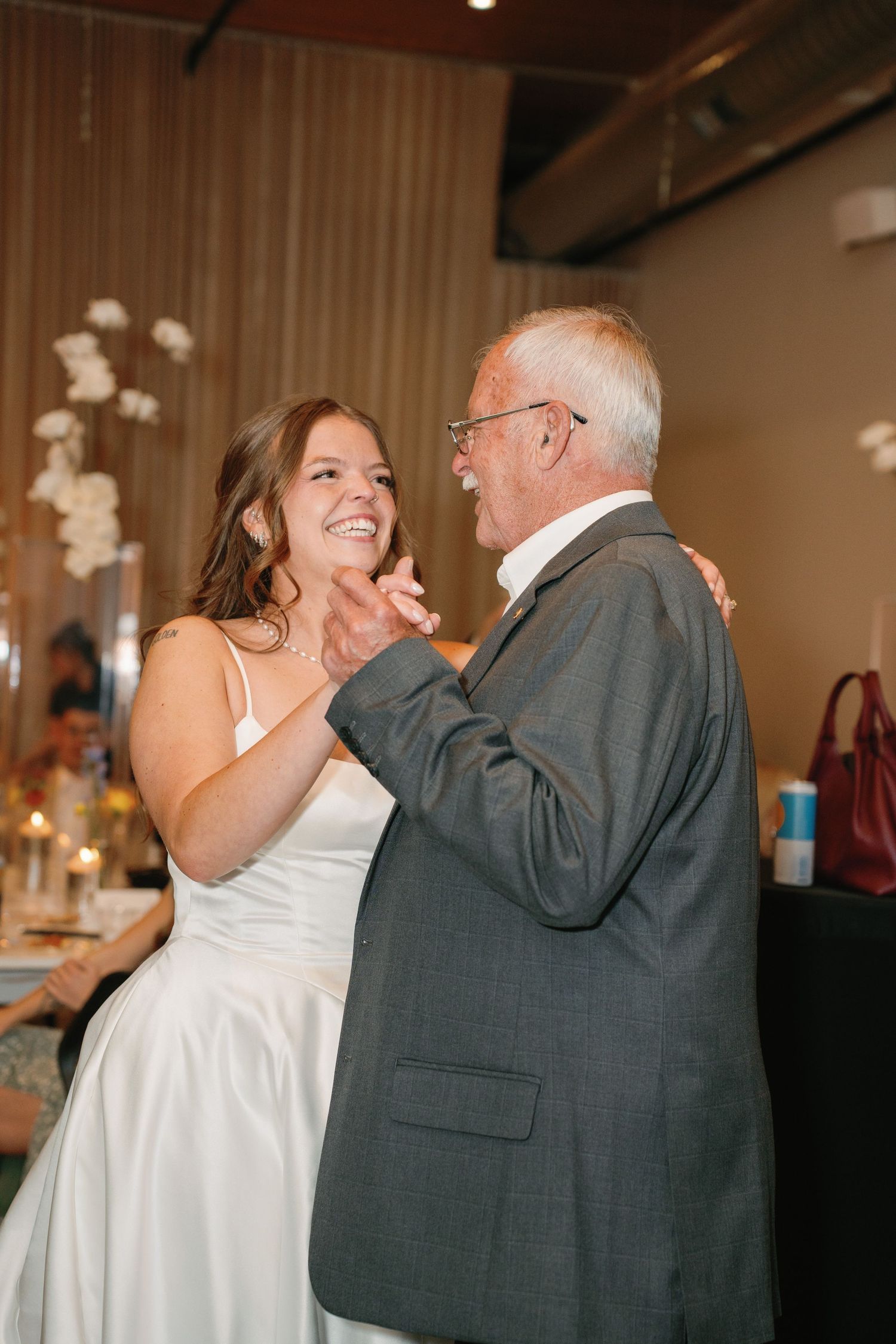 Bride in white satin dress shares joyful father-daughter dance at elegant indoor wedding reception.