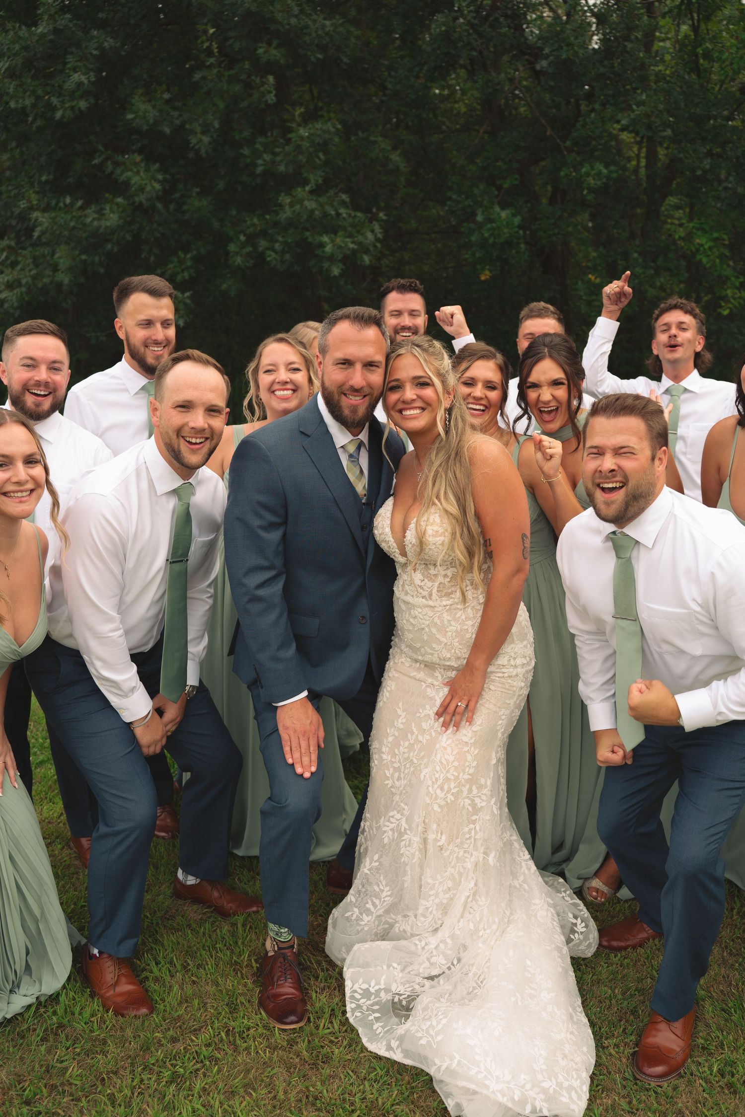 Wedding party members strike energetic poses together outdoors in sage green and navy attire.