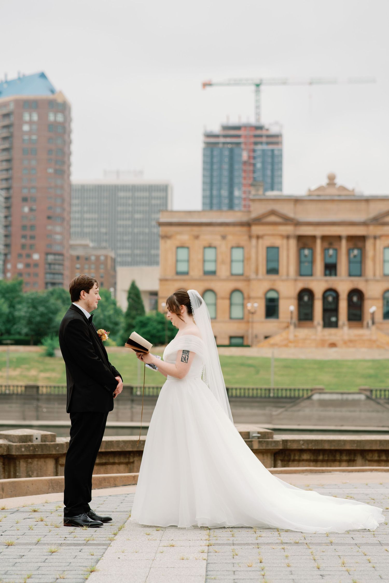 Bride and groom exchange vows outdoors with urban skyline and historic building backdrop on their wedding day.