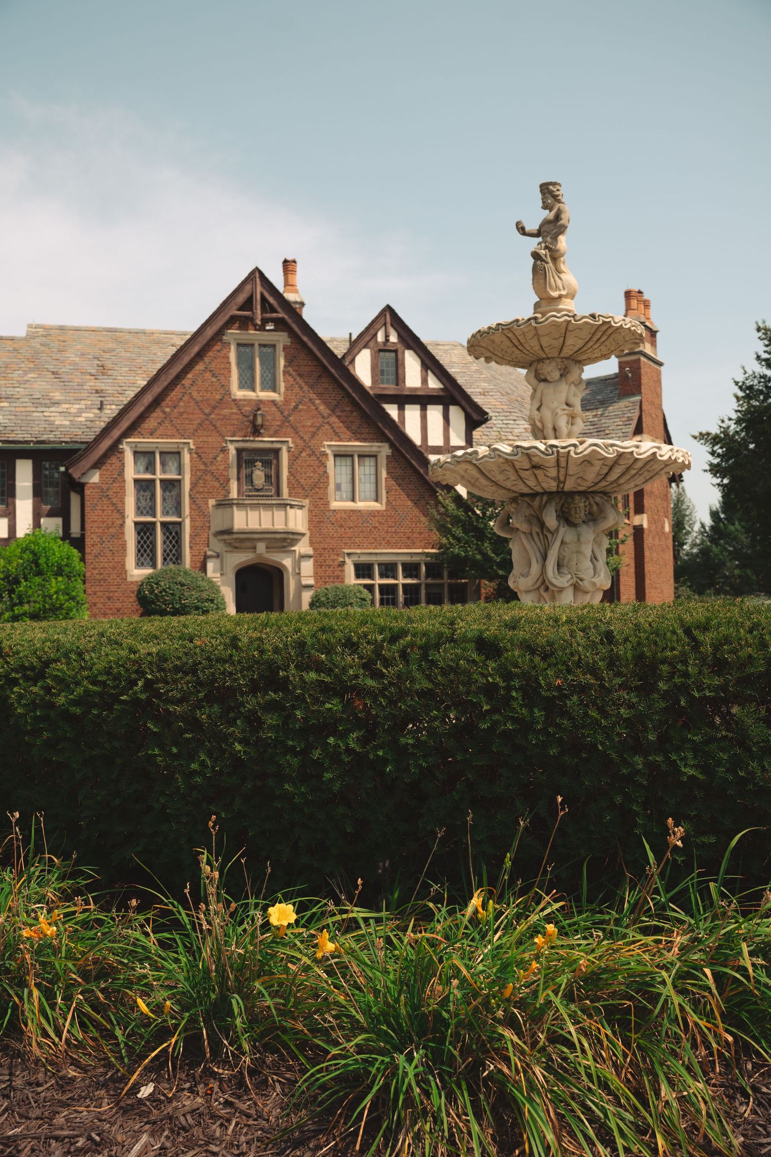 Ornate brick mansion with Tudor-style architecture features decorative statues and a well-maintained hedge garden in the foreground.