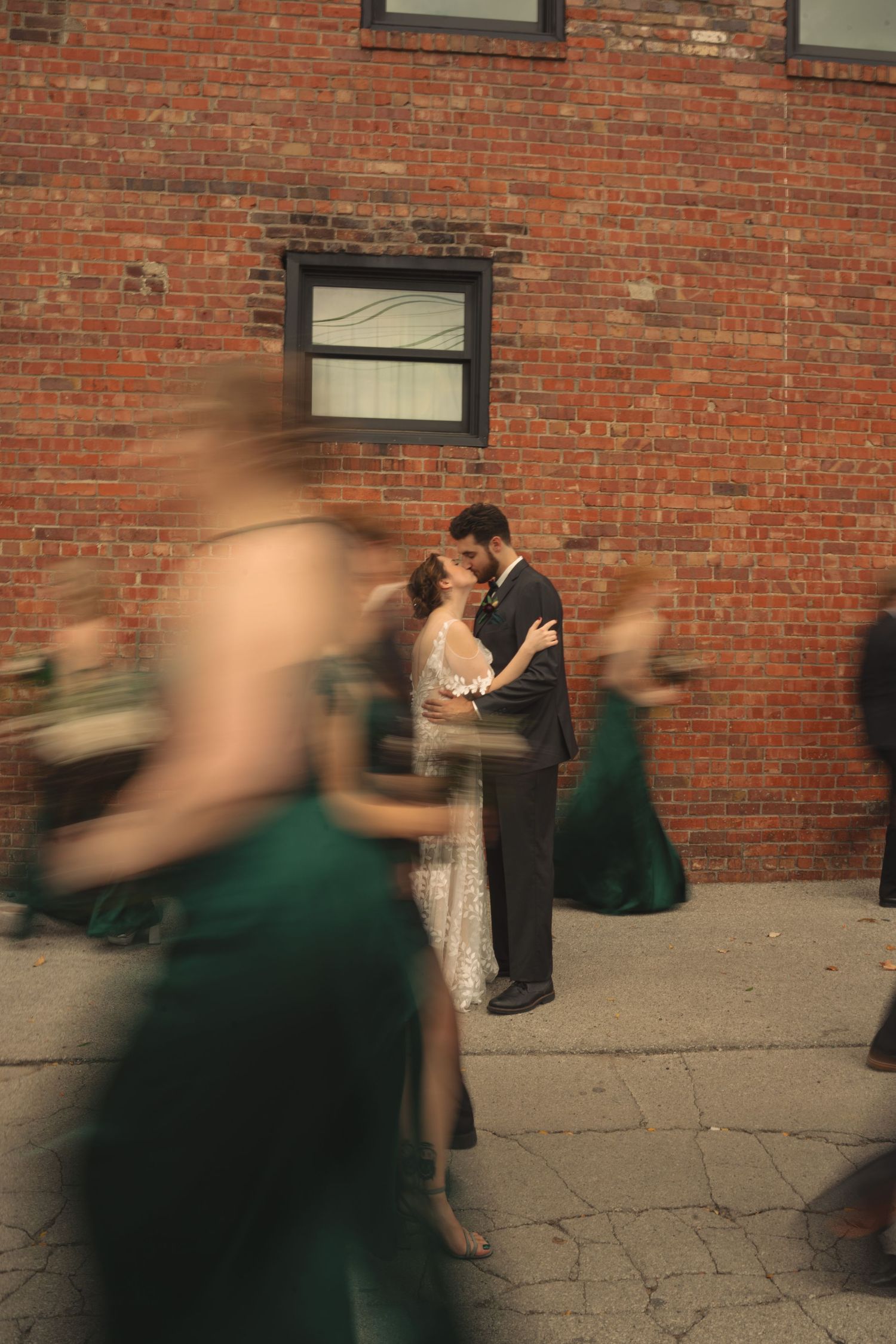 Wedding couple embraces against brick wall as guests create motion blur in foreground.