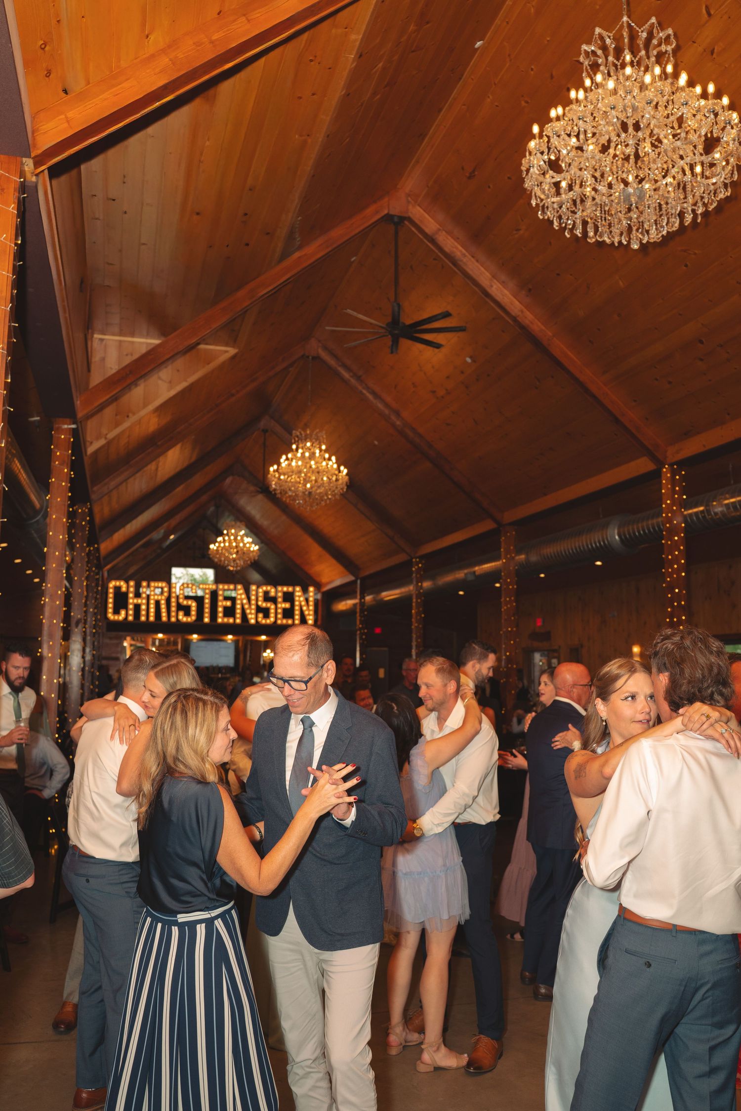 Wedding guests slow dancing under elegant chandeliers in a wooden-beamed reception hall.