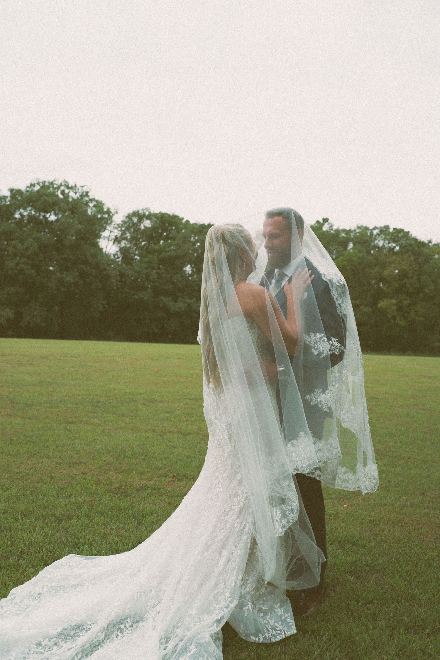A couple embraces under flowing lace veils in an open field during their wedding.