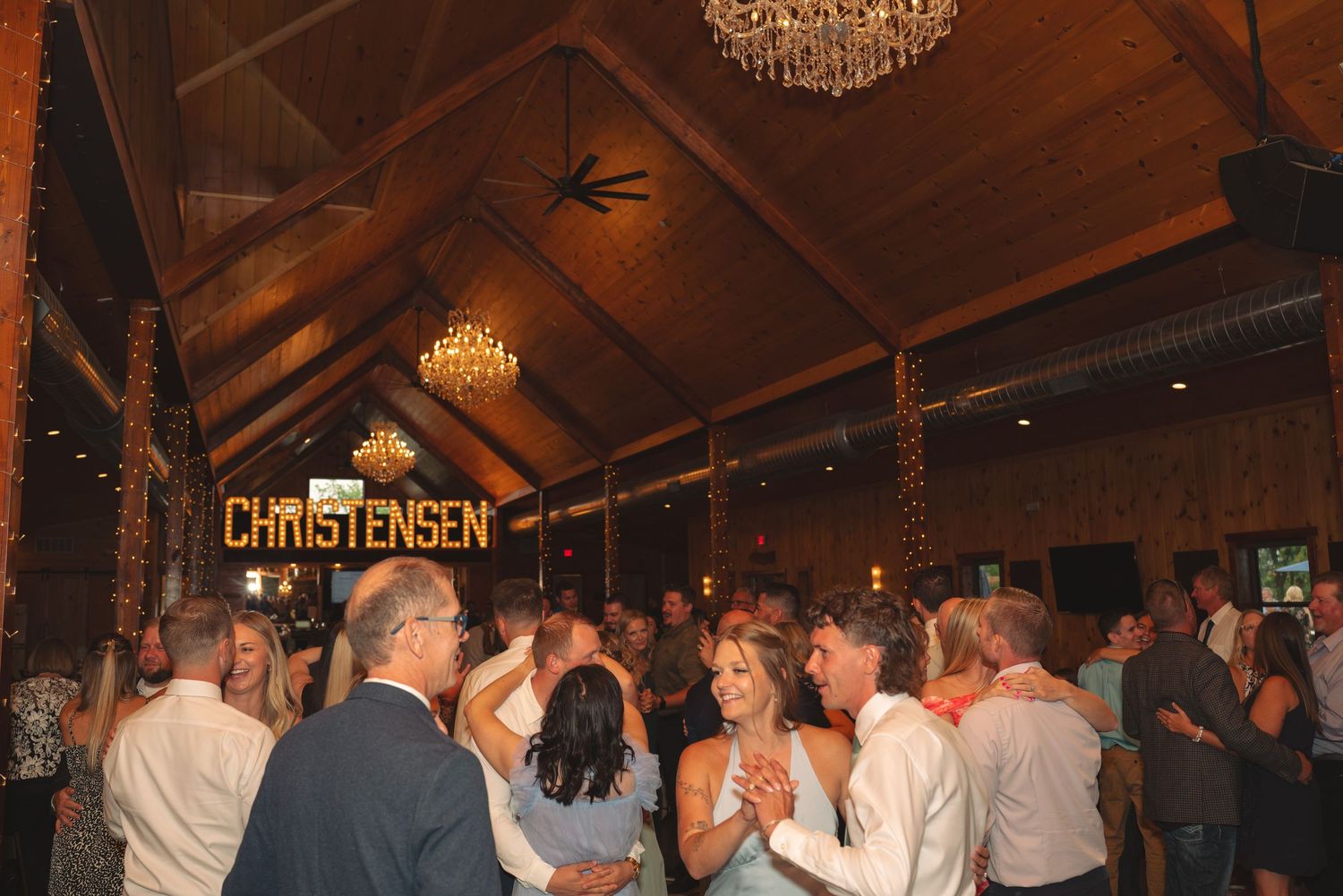 Wedding celebration with guests dancing under crystal chandeliers in a rustic barn setting.