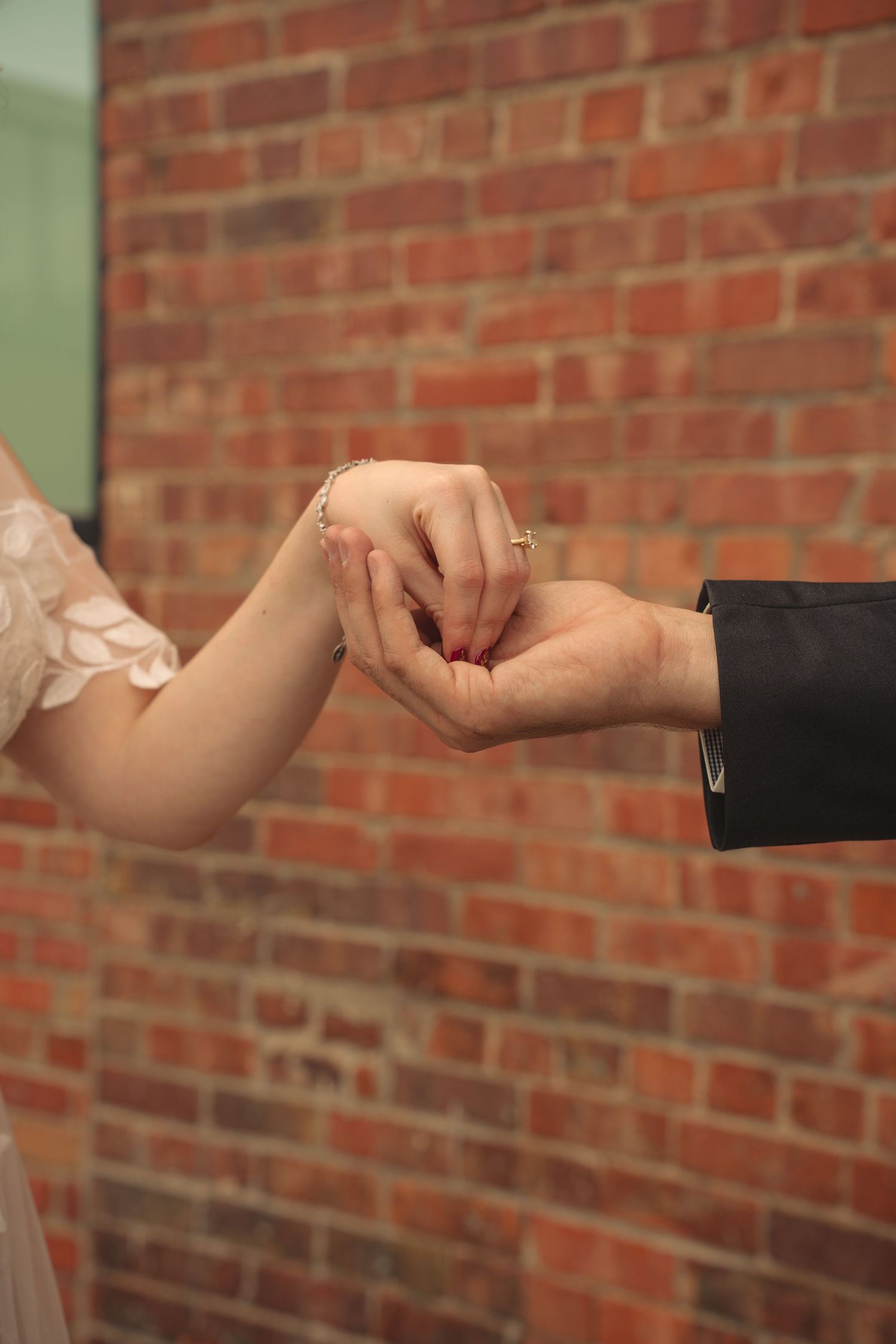Close-up of bride and groom's hands touching tenderly against brick wall background.