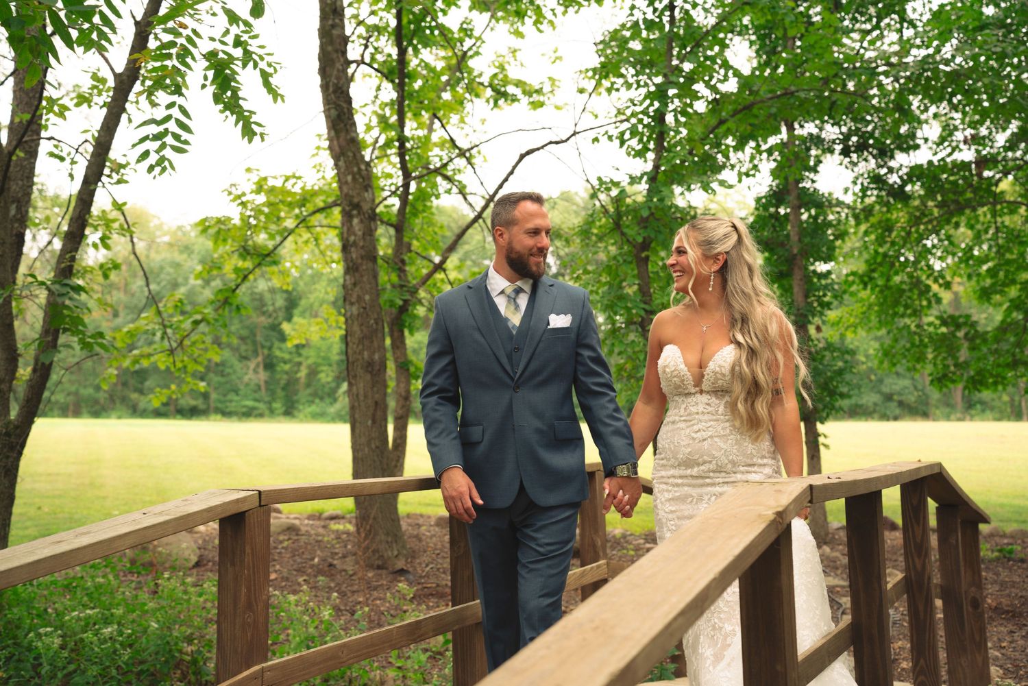 Romantic wedding couple walking along a rustic wooden bridge surrounded by lush green trees.
